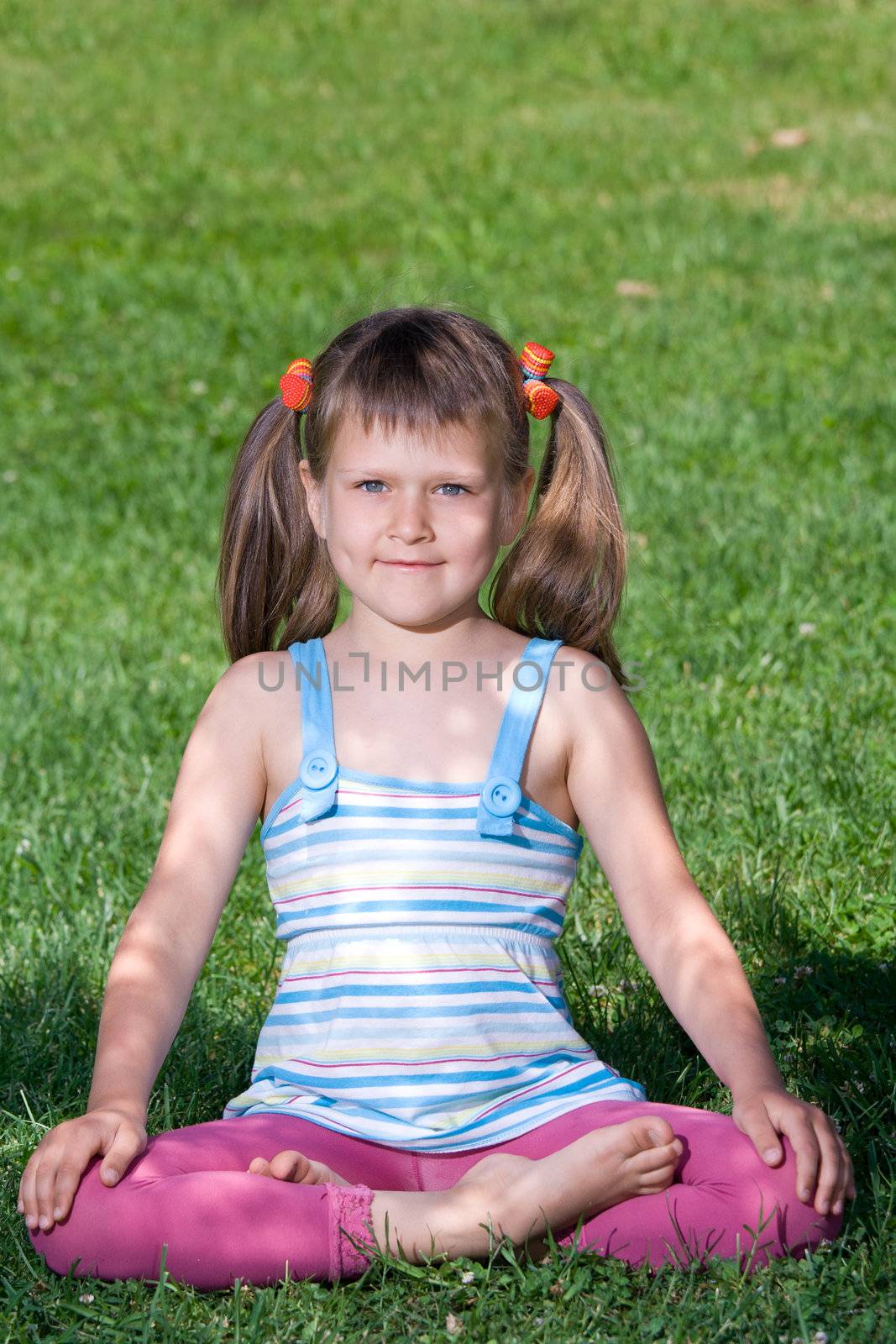 Smiling happy little cute girl who is sitting and meditating in yoga lotus asana under tree shadow on the green grass