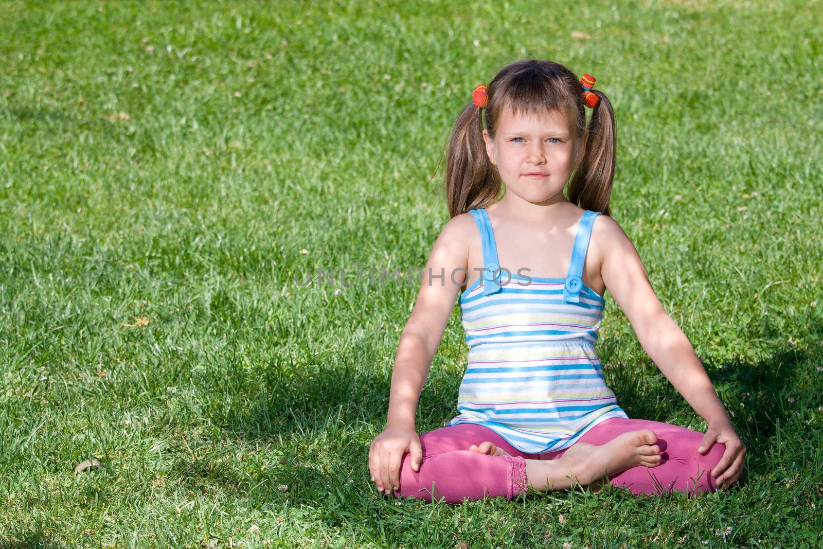 Cute little girl is sitting in yoga lotus asana under tree shadow on the green grass