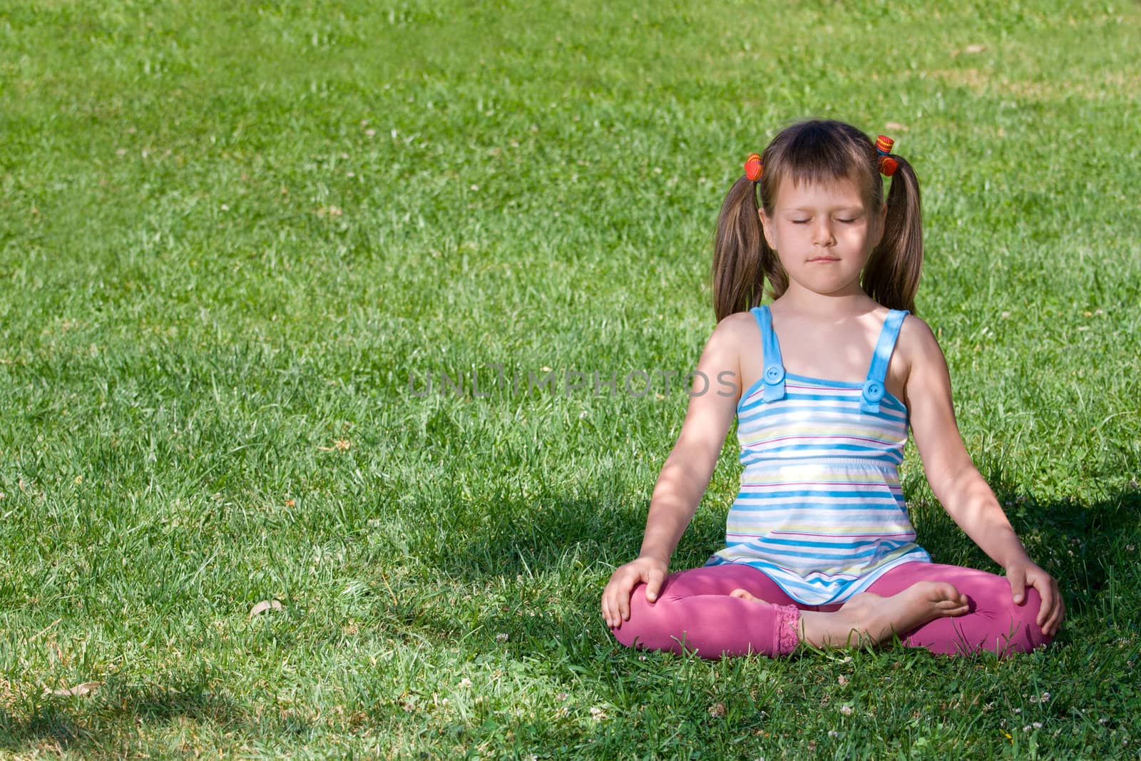 Healthy lifestyle. Little cute girl who is sitting and meditating in asana under tree shadow with closed eyes on the green grass