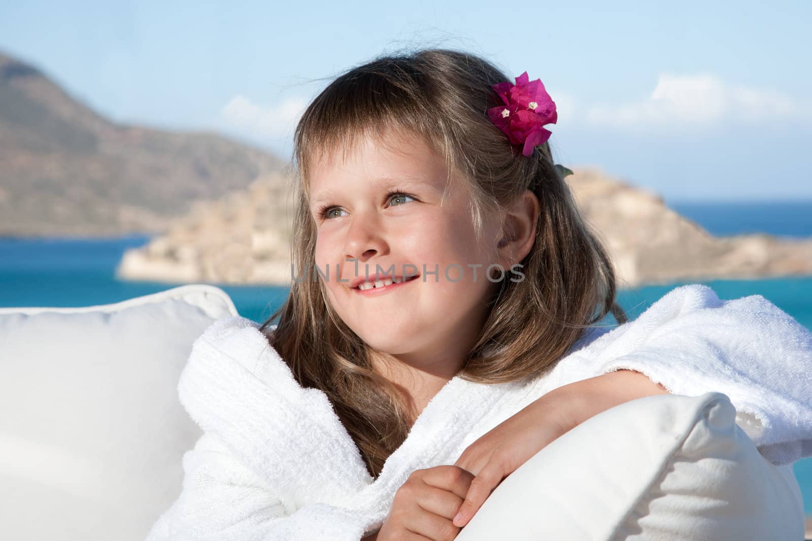 Portrait of happy smiling little girl with flowered hair in white bathrobe relaxing on terrace divan on the sea background