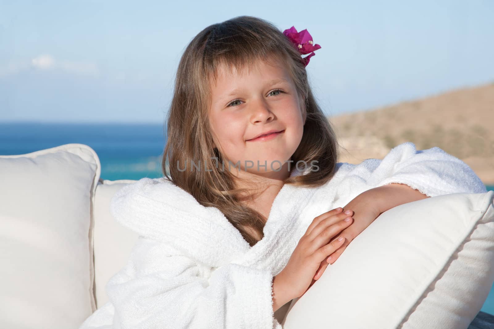 Portrait of happy smiling little girl with flowered hair in white bathrobe relaxing on terrace divan and looking at camera on the sea background