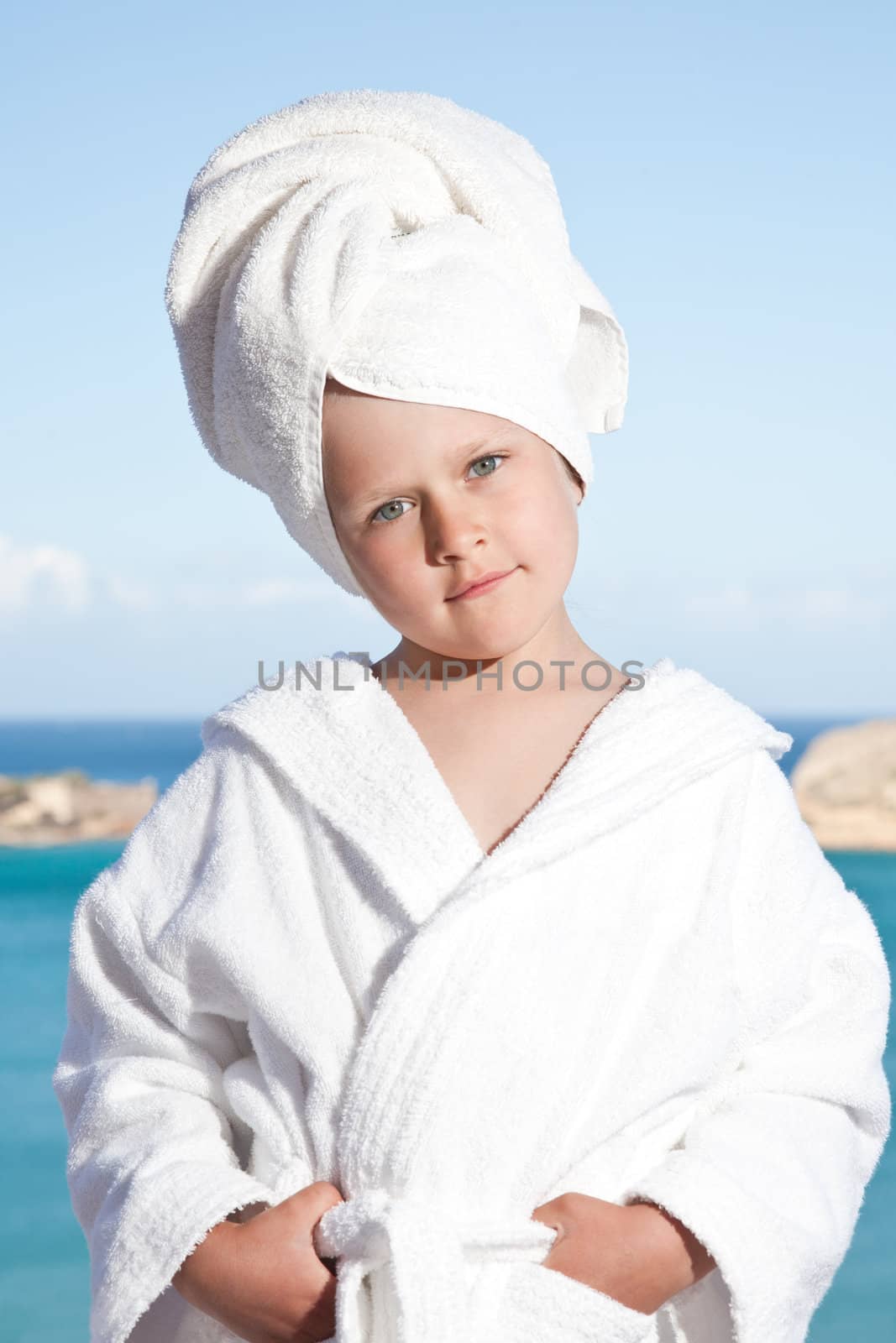 Portrait of happy smiling little girl with towel on the head in white bathrobe relaxing on terrace on the sea background