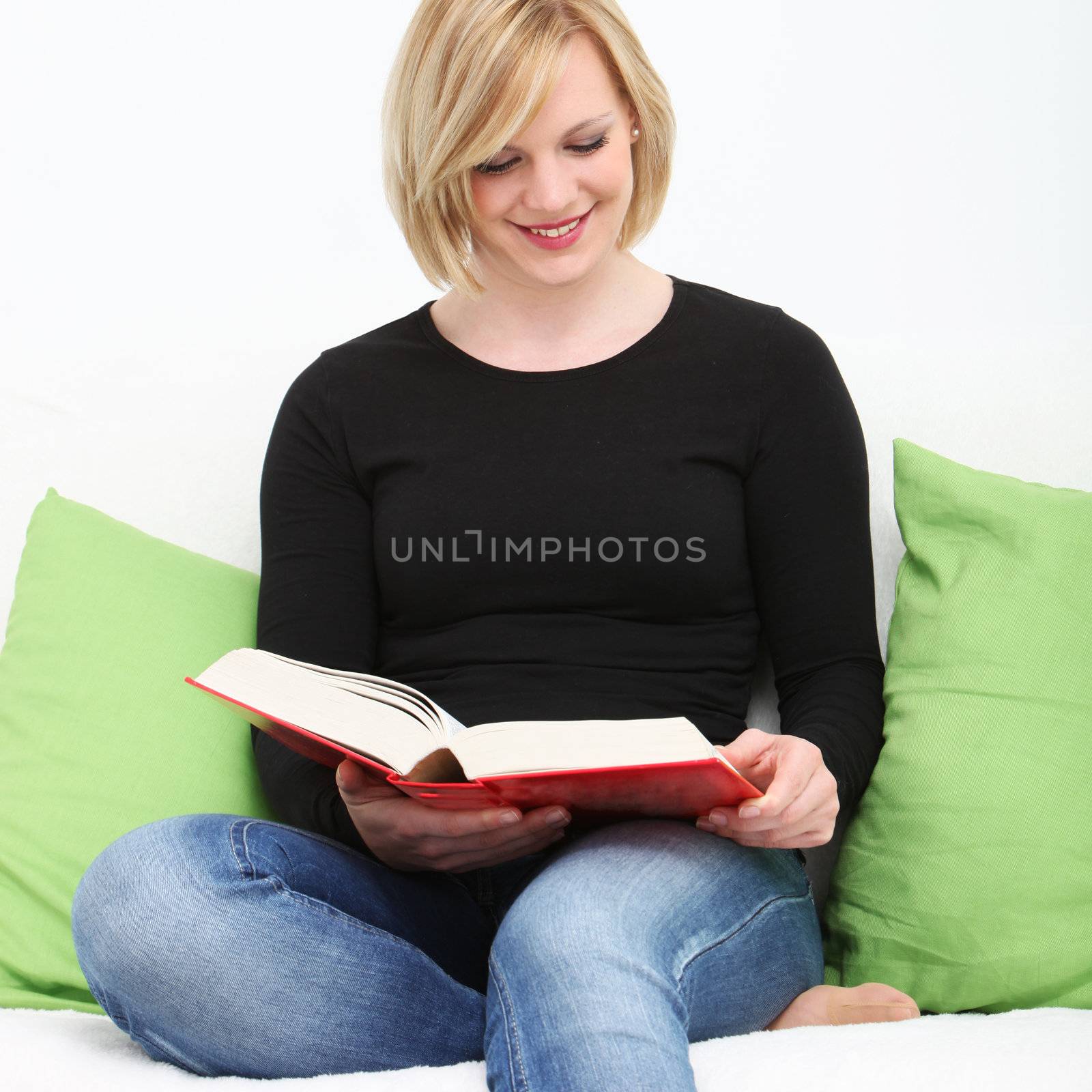 Closeup portrait of a young smiling woman sitting on a couch in jeans with her bare foot curled up under her reading a book