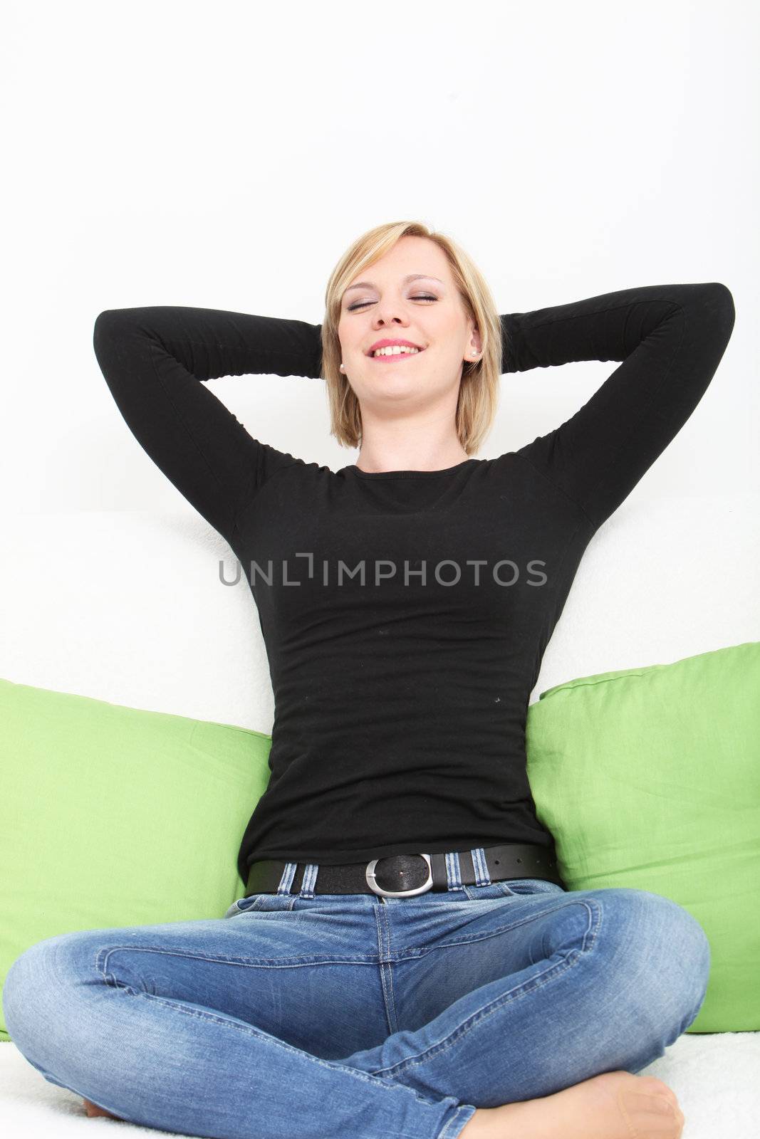 Attractive casual young woman in jeans and bare feet sitting back relaxing on a couch with her hands behind her head and a look of blissful pleasure on her face