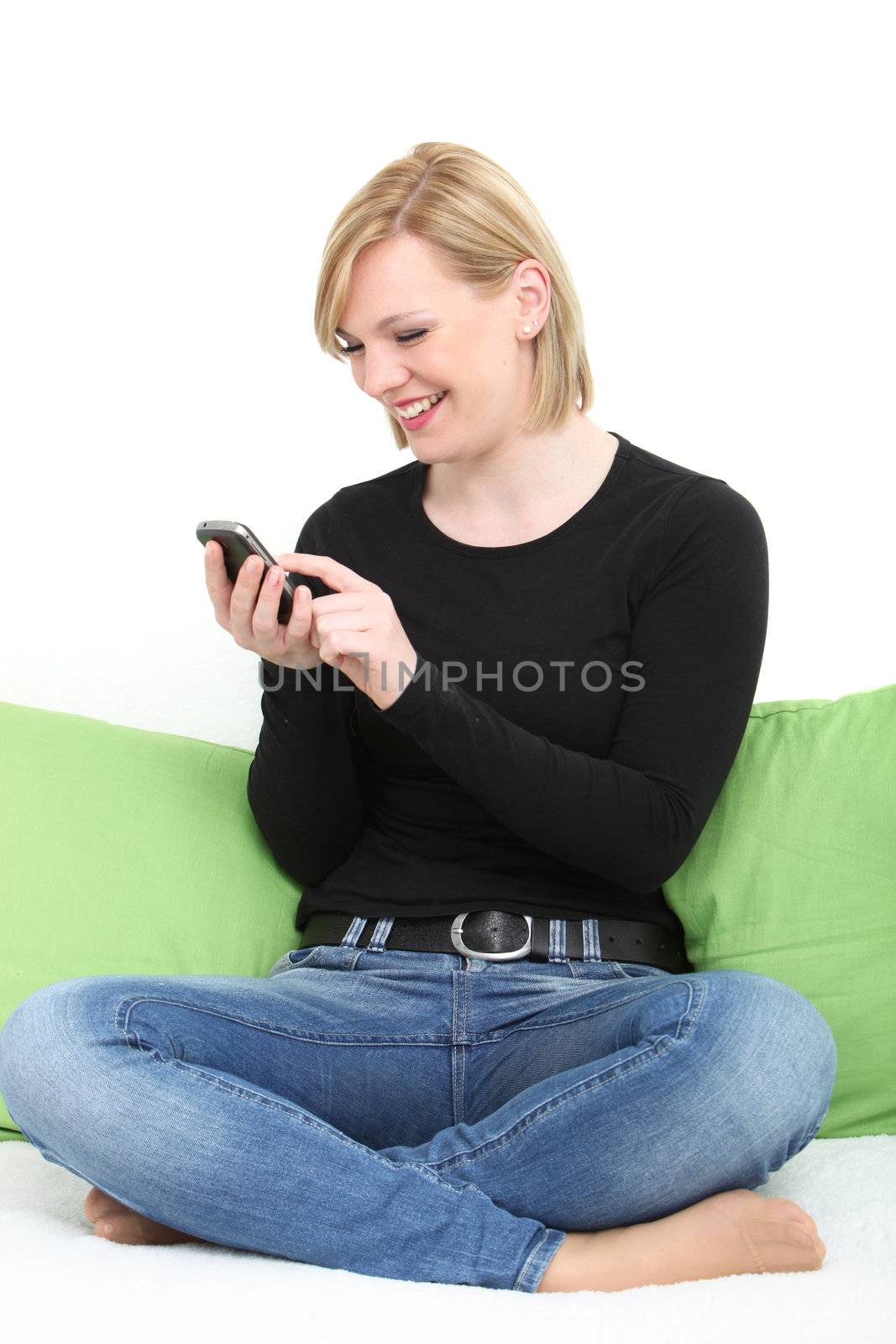 Happy young woman sitting cross legged on a couch barefoot in jeans texting on her mobile phone using her finger to type on the touchscreen