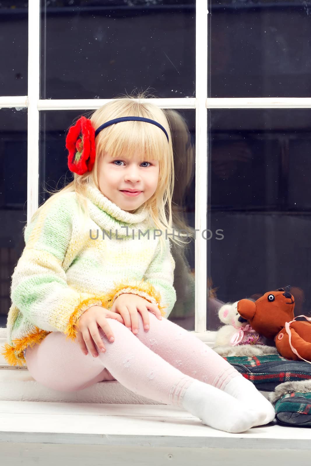 beautiful little girl blonde in a knit sweater sitting beside toys in the window and looks into the camera