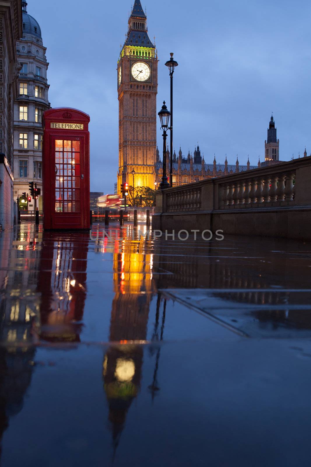 Red public telephone box and illuminated clock on Big Ben tower of Westminster Palace in twilight with reflection on wet footway, Great Britain