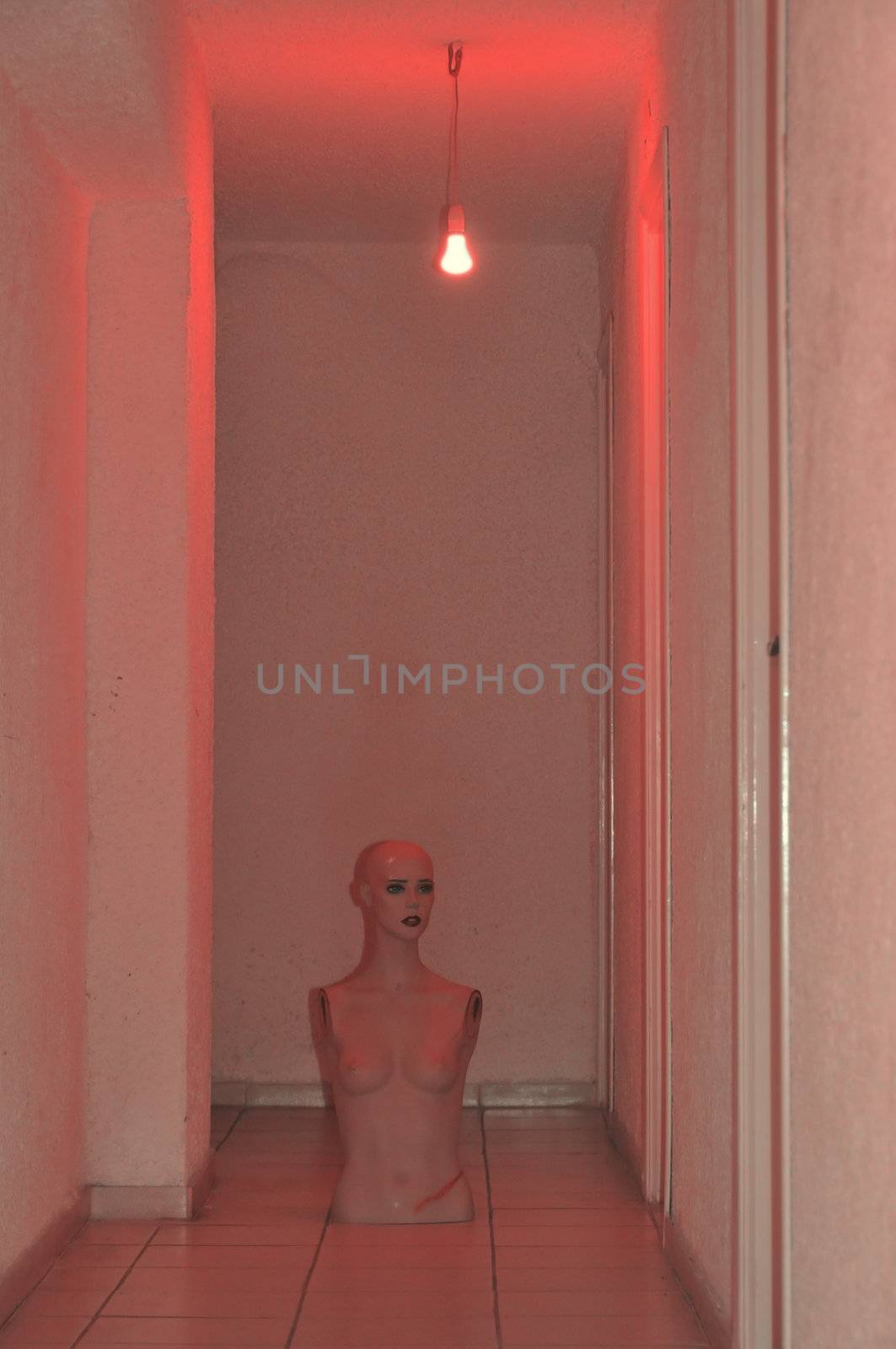 Broken mannequin doll under red light in underground corridor.