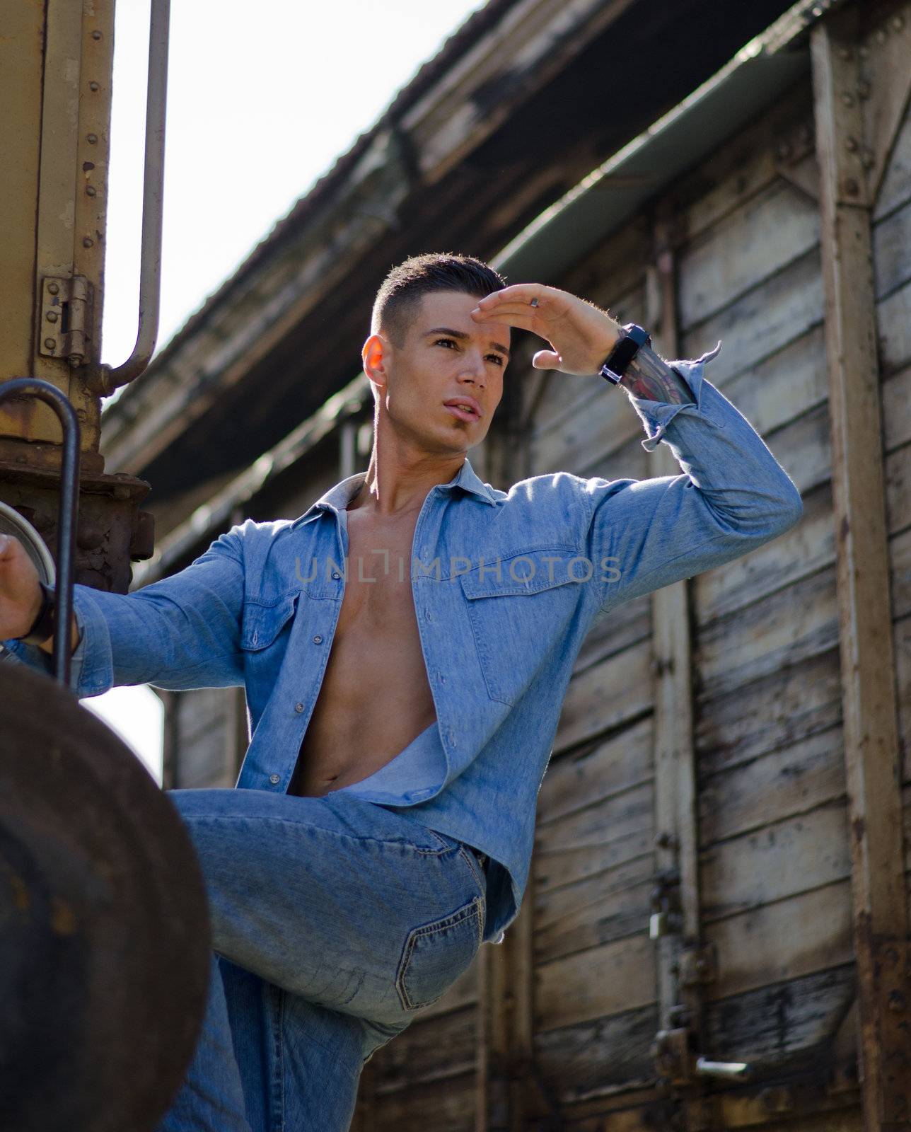 Fit guy with shirt open looking in the distance hanging from old train