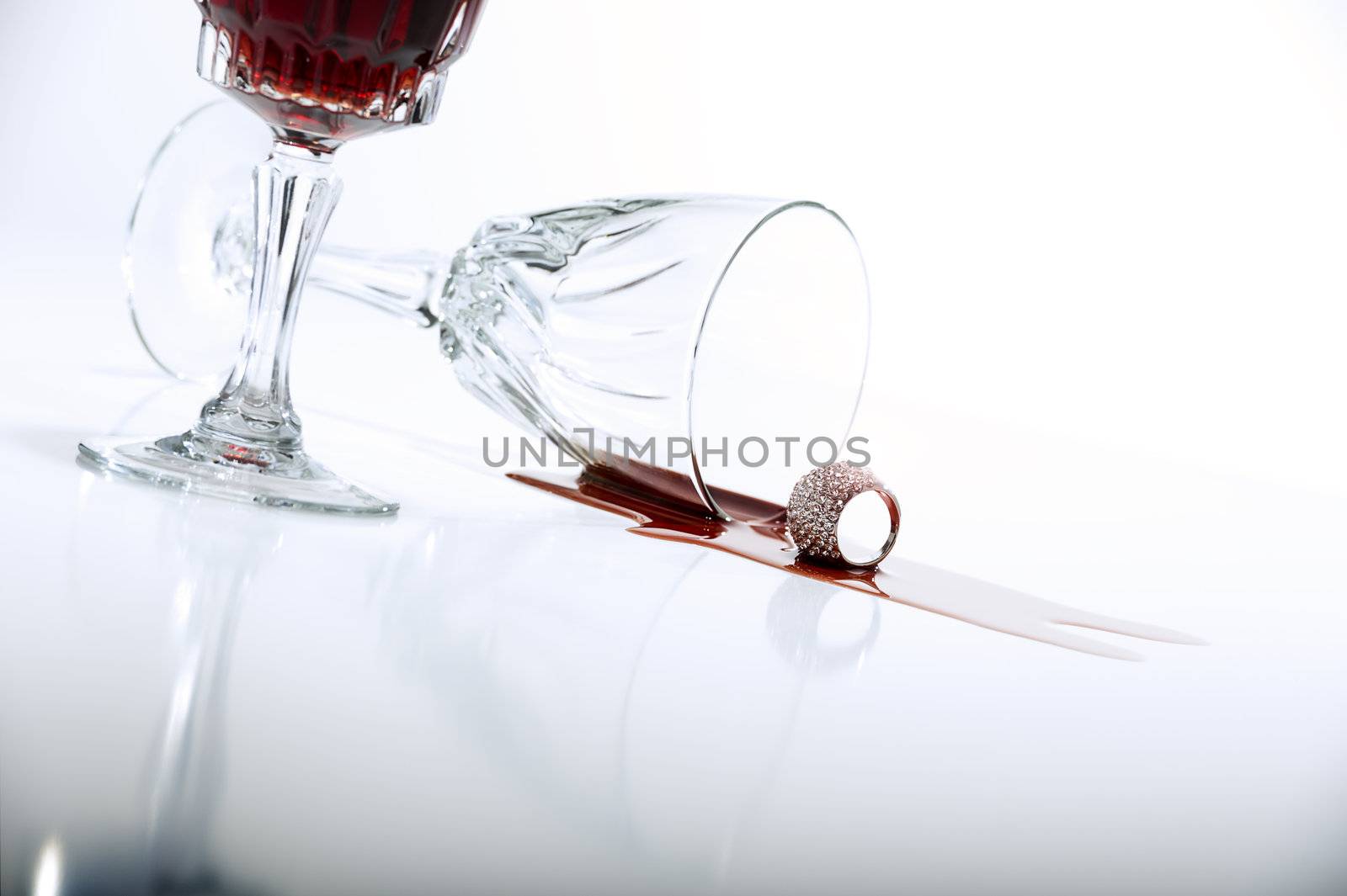 Low angle view across a reflective white countertop of a pool of spilled red wine from crystal wineglass lying on its side alongside a full cropped upright glass with copyspace