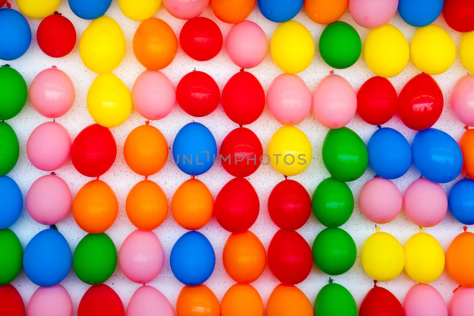 A white wall with colorful balloons attached. Photo is of a boardwalk arcade game where you throw darts and try to pop balloons. Only the wall of balloons is shown.