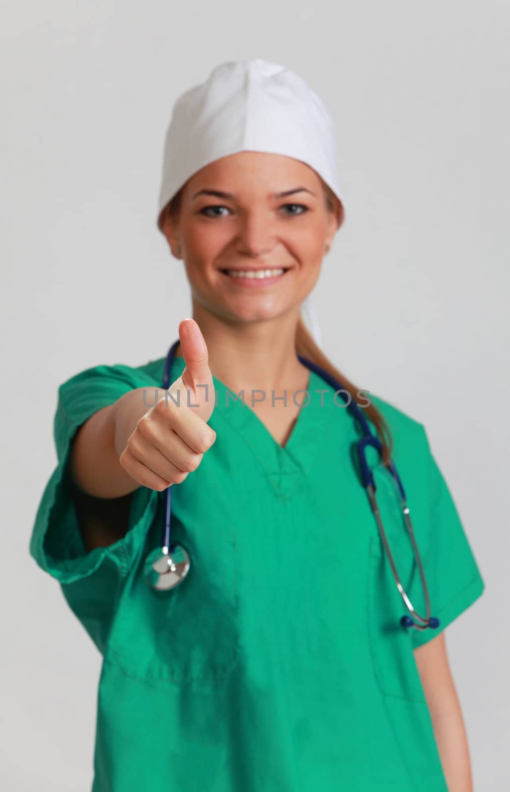 Portrait of a smiling young woman doctor with her right thumb up, against a gray background. Selective focus on the thumb.