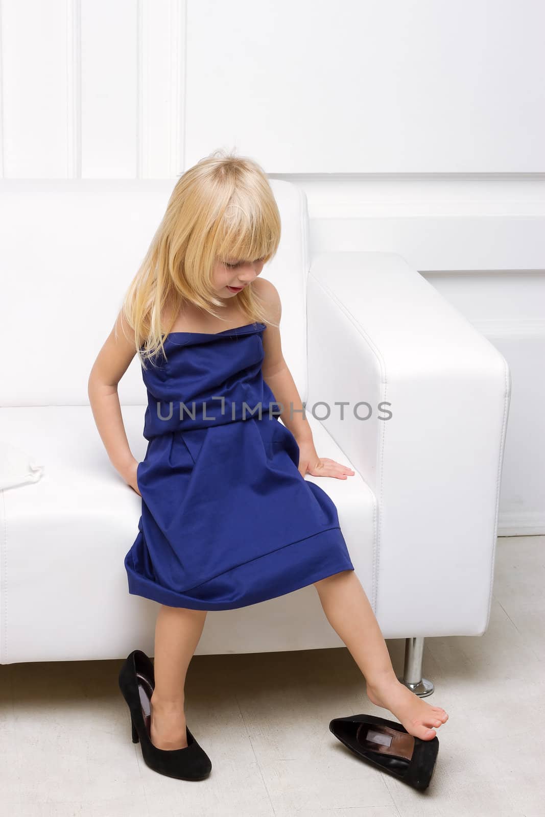 Little girl 3 years old standing near the white sofa in her mother's dress shoes and high heels