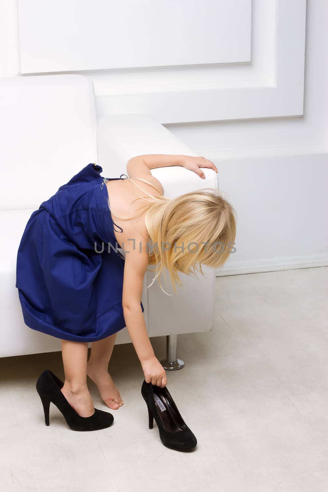Little girl 3 years old standing near the white sofa in her mother's dress shoes and high heels