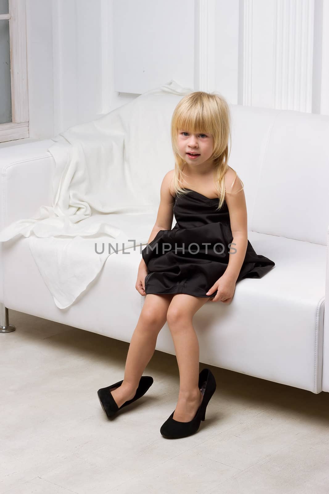 Girl 3 years old in her mother's high heels sitting on a white sofa