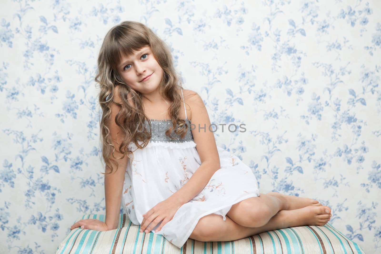 Portrait of sincere cheerful tender young blond girl child with grey eyes and wavy long hair in relaxed sitting posture looking at camera