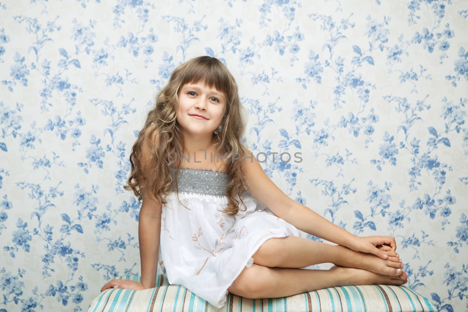 Portrait of sincere cheerful tender young blond girl child with grey eyes and wavy long hair in relaxed sitting posture looking at camera