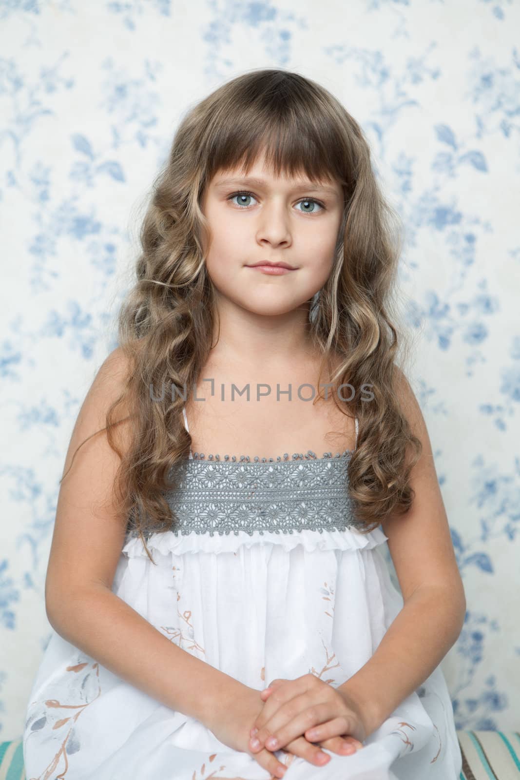 Portrait of sincere cheerful tender young blond girl child with grey eyes and wavy long hair in sitting posture looking at camera