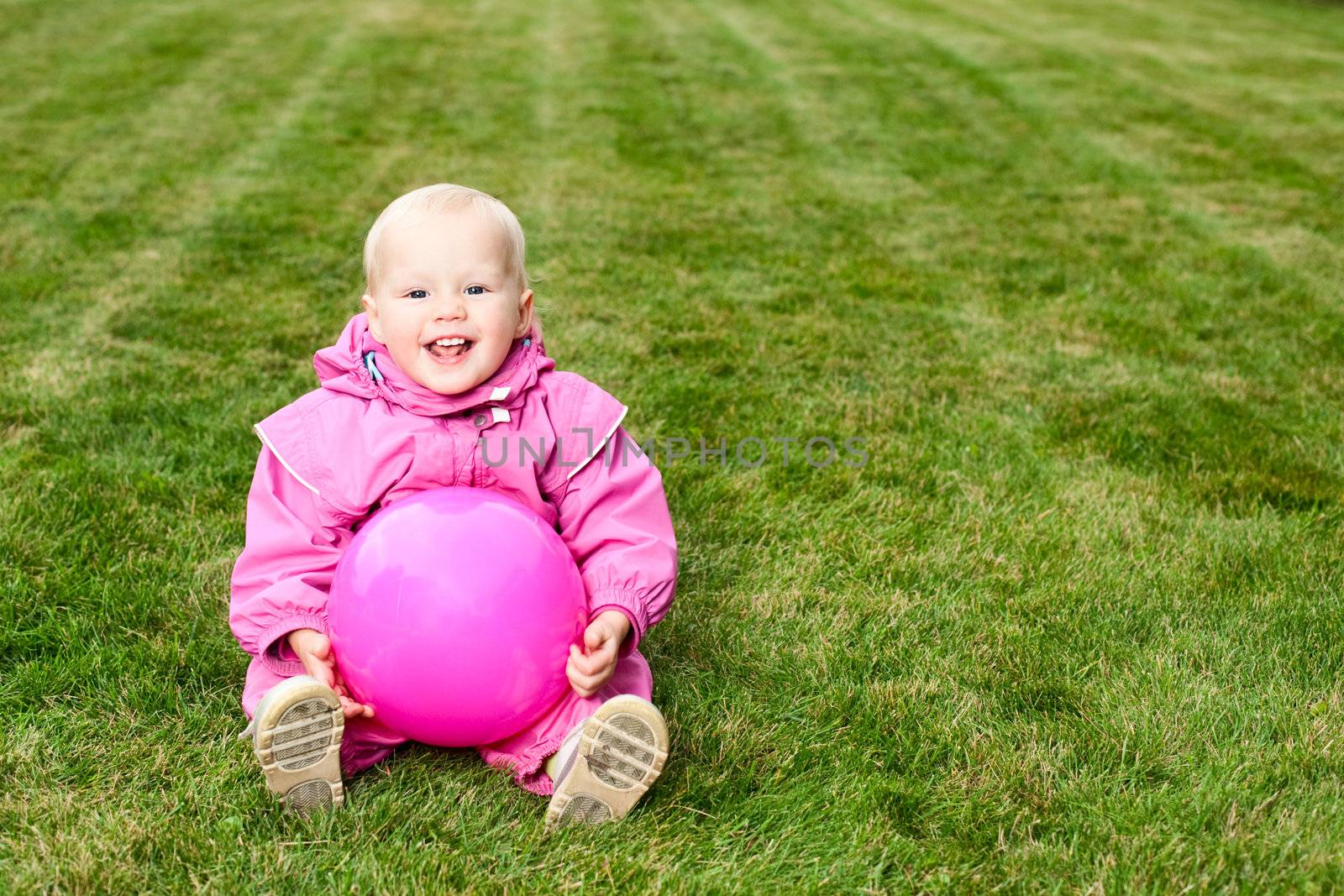 Cute little baby girl wearing pink suit sitting with pink ball on lawn