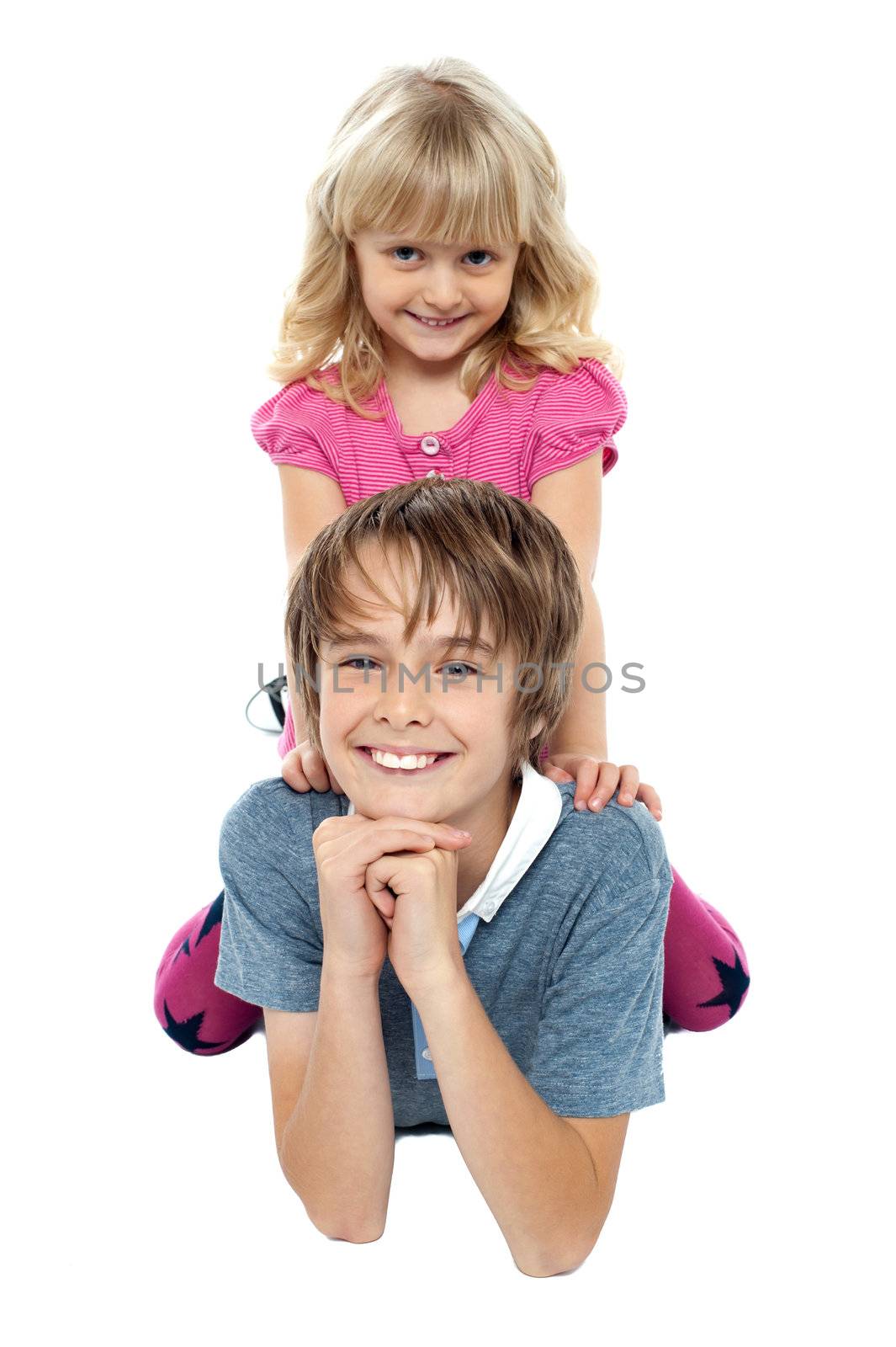 Adorable sister sitting on her brothers back while he lies on studio floor with cheeky expression.