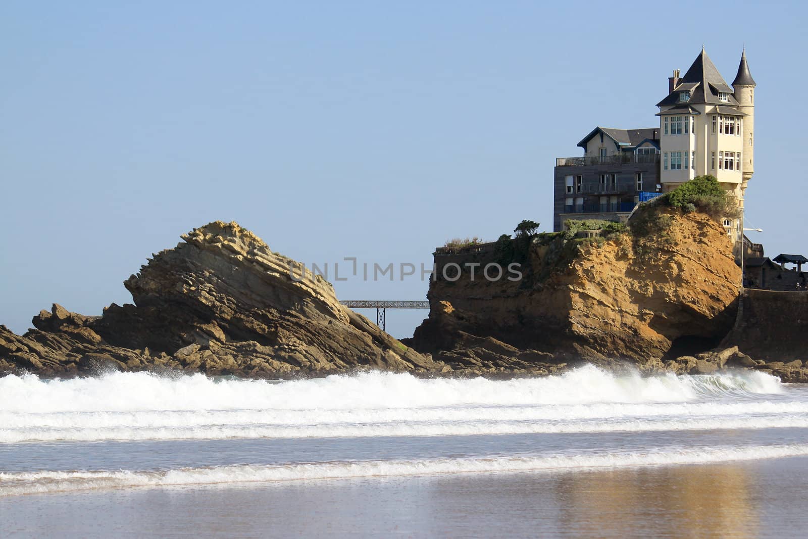 An old mansion perched on a rock in the ocean near a beach on a blue sky background