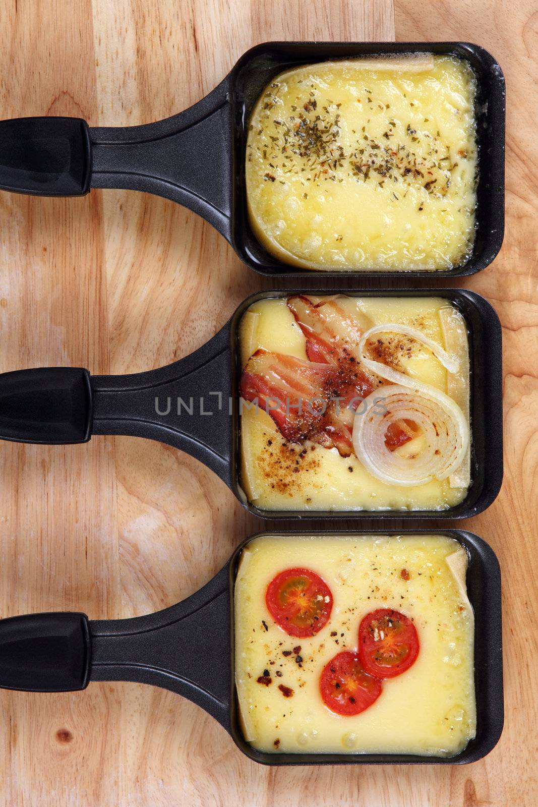 Photo of a three Raclette trays with melted cheese, herbs, cherry tomato, bacon and onion.