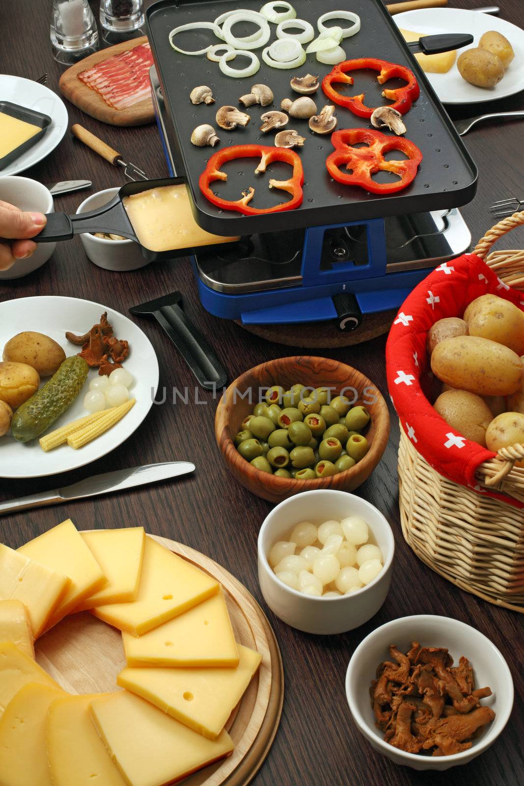 Photo of a table full of items for a traditional Raclette dinner, including slices of cheese, white onions, potatoes, olives, mushrooms, and the grill with the utensils.
