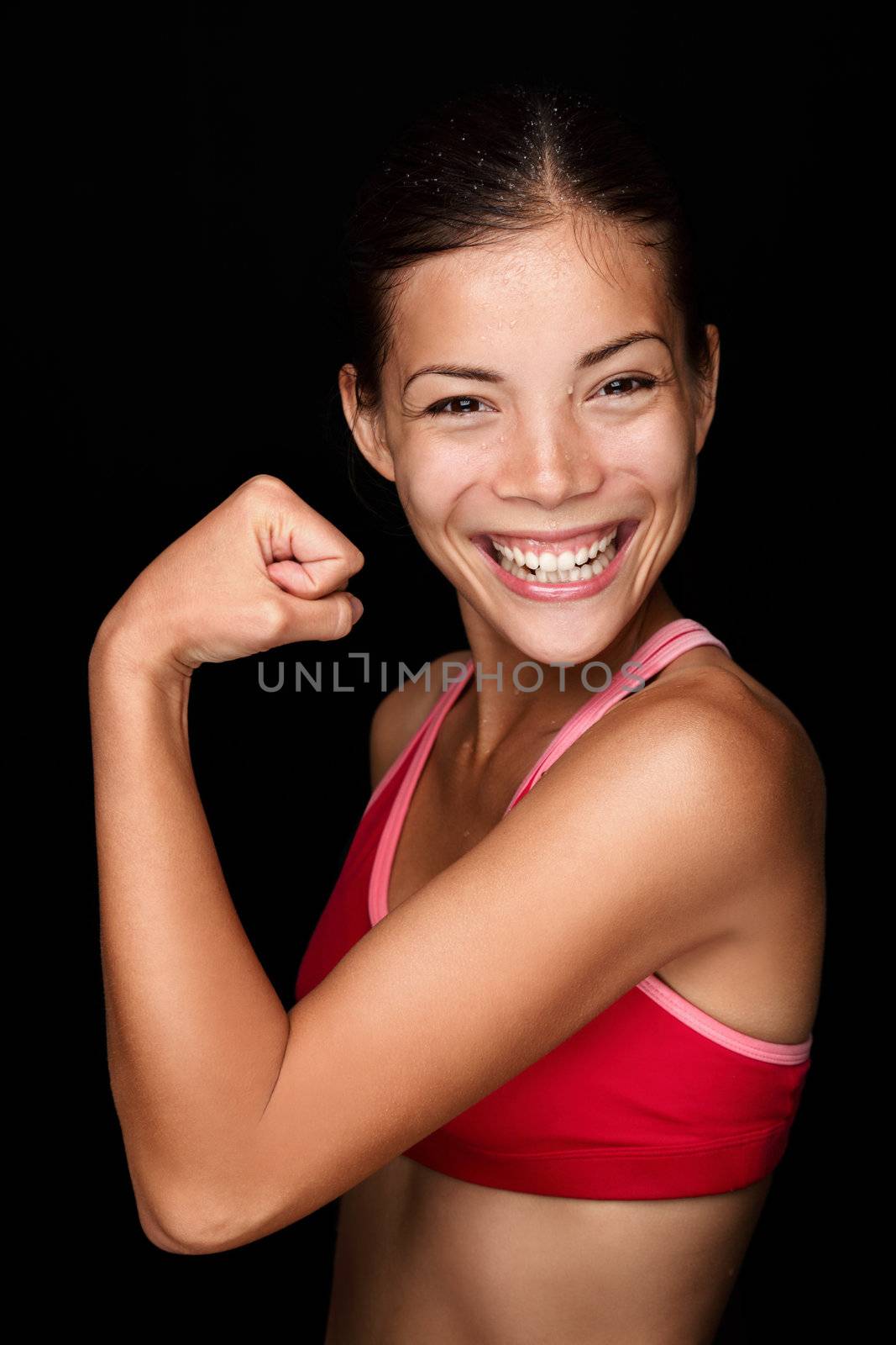 Playful young female Asian athlete flexing her arm with a mischievous smile to display her arm muscles. Fun upper body portrait on a black background