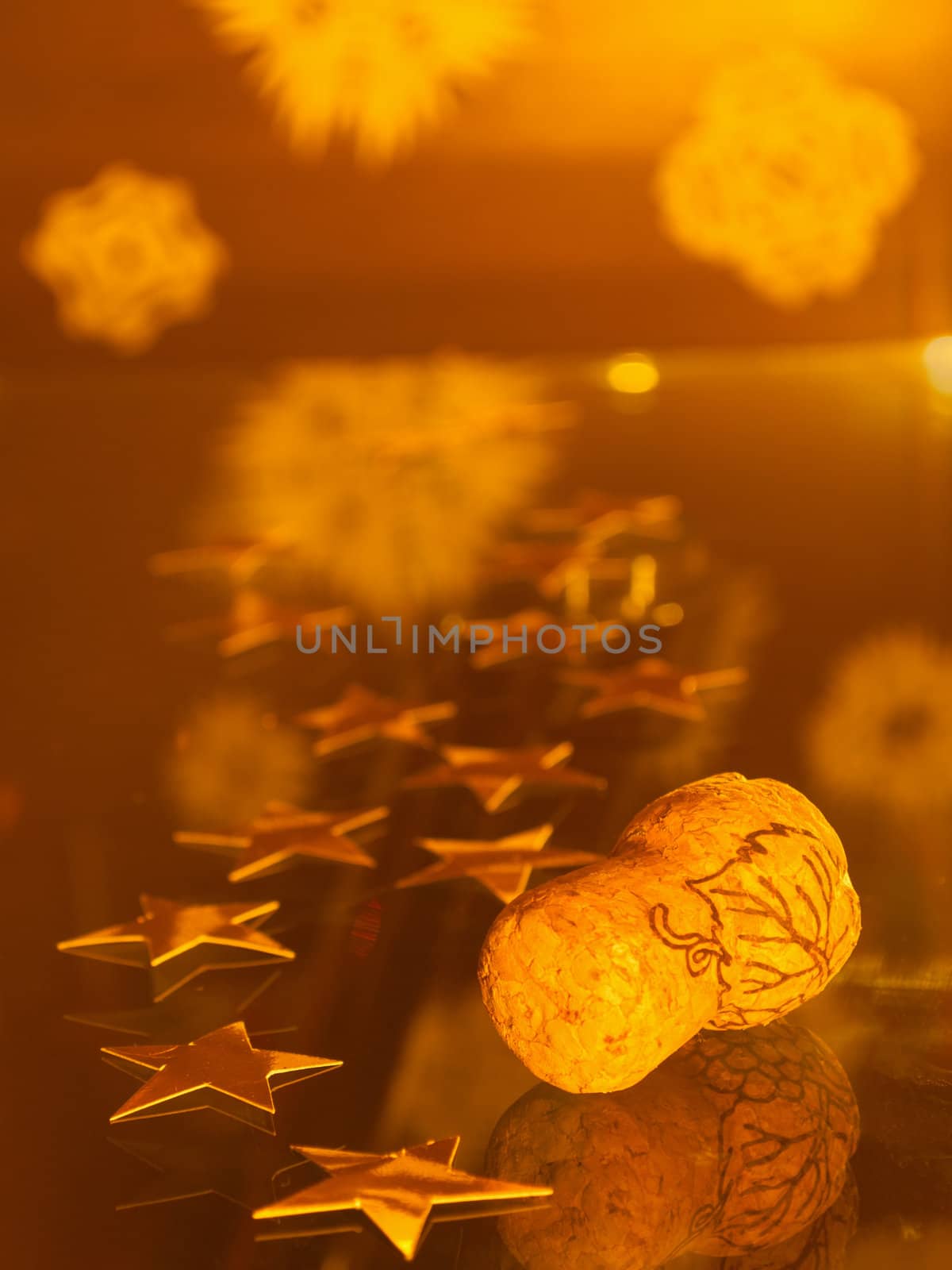 Champagne cork and golden stars lay on glass surface with paper snowflakes behind; focus on cork