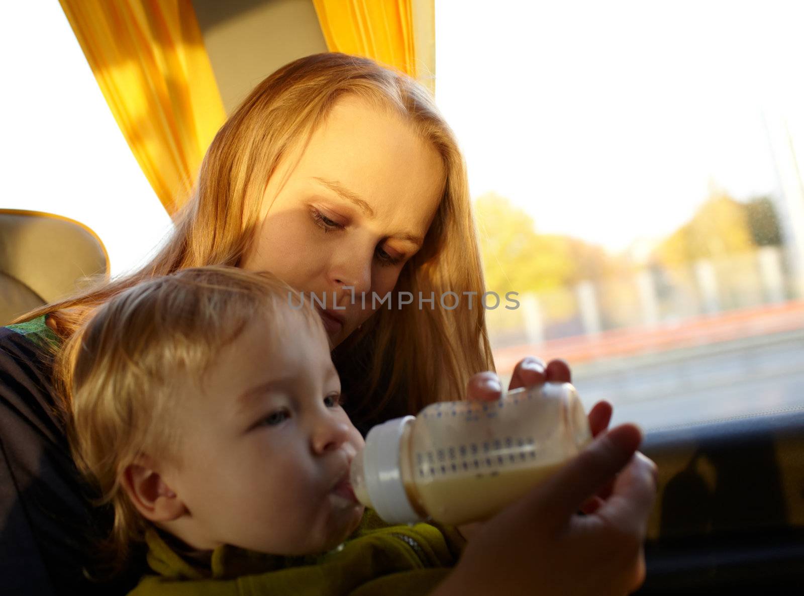 Young mother is feeding her two year old son with milk, while they are travelling by intercity bus. Close up portraits with natural light.