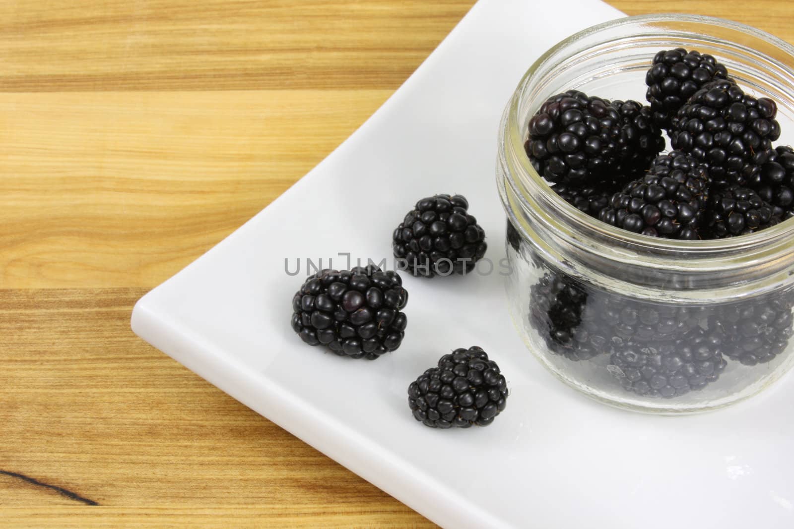 Blackberries in a jar, on a white plate sitting on a wooden counter top.
