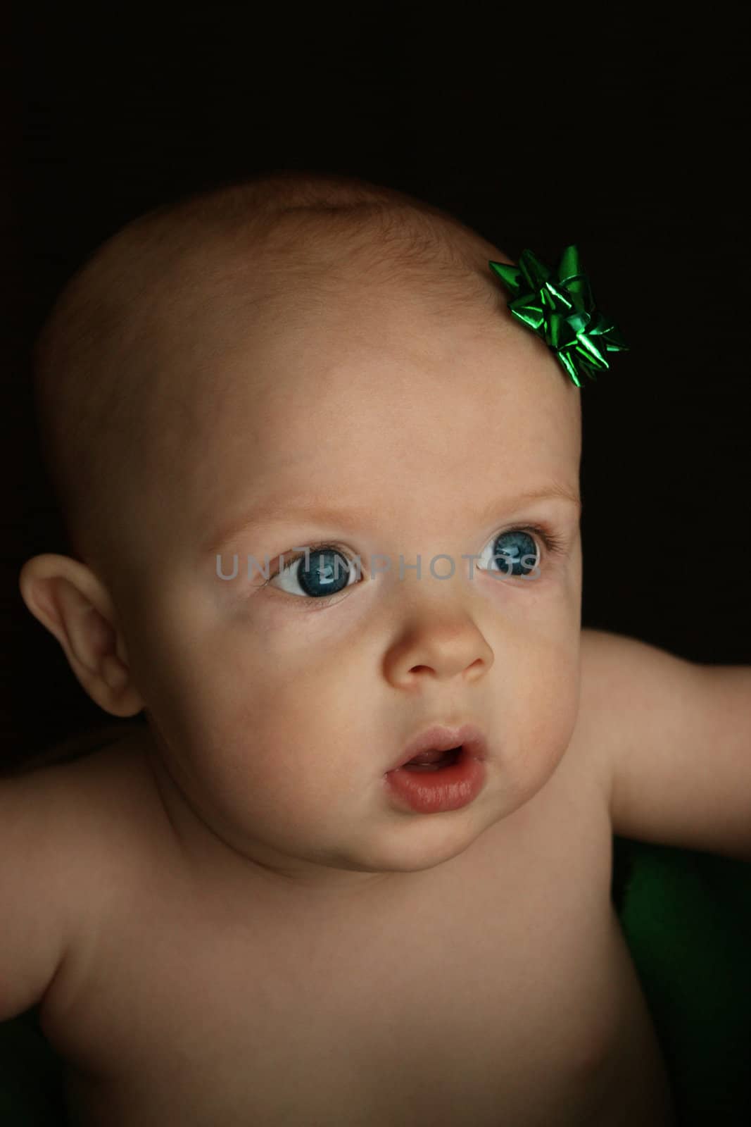 A close up of an infant with a green Christmas gift bow on his head, sitting in a wicker basket,  on a black background.