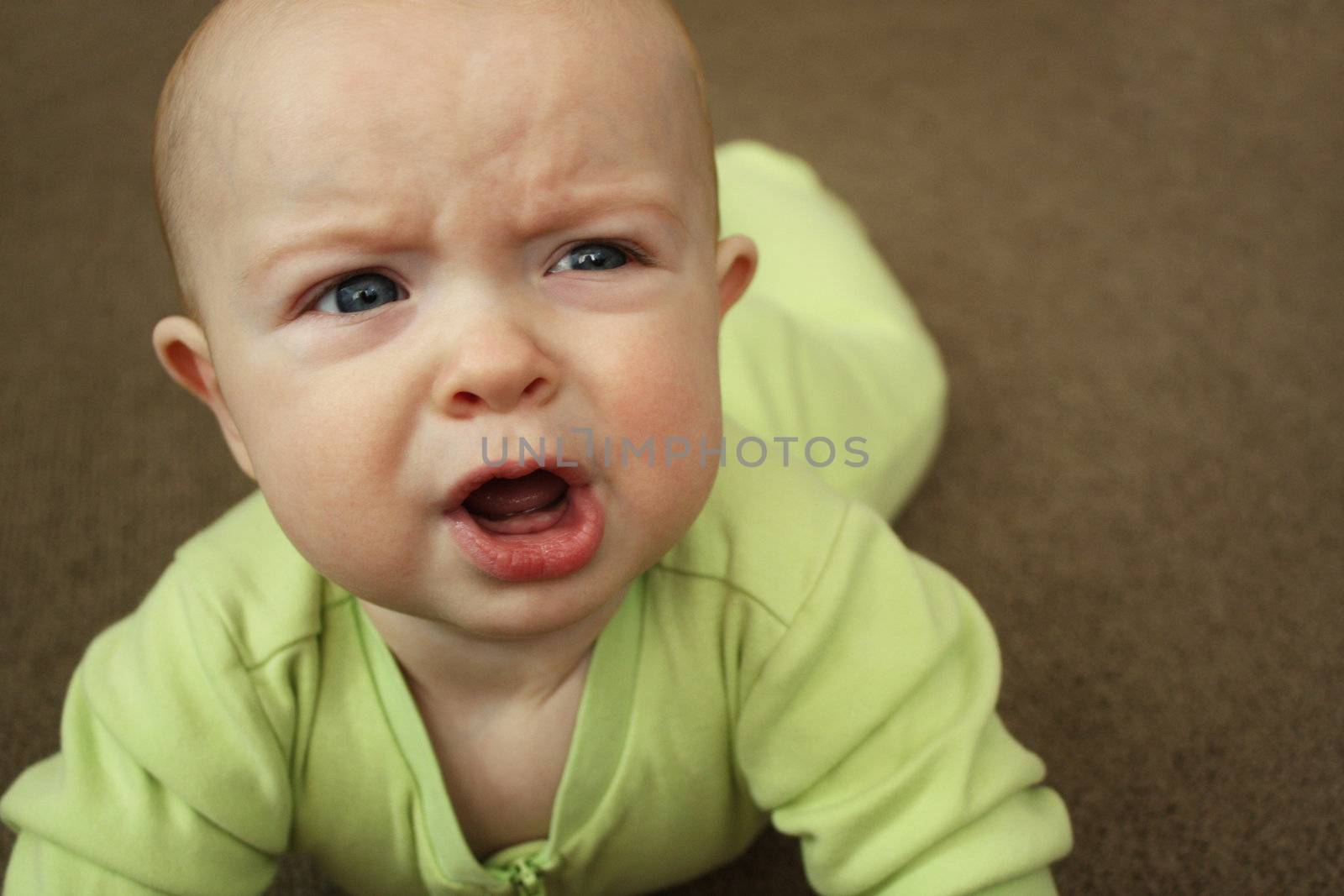 A close-up of a baby in a light green outfit on a brown carpet, crying.