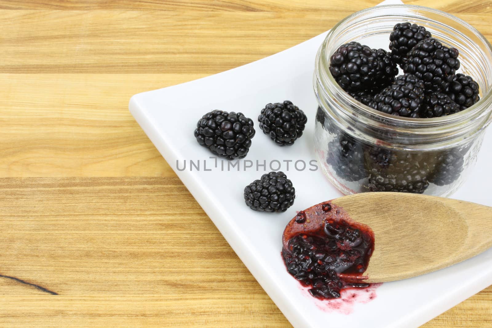 Blackberries in a jar, with blackberry jam on a wooden spoon, all on a white plate sitting on a wooden countertop.