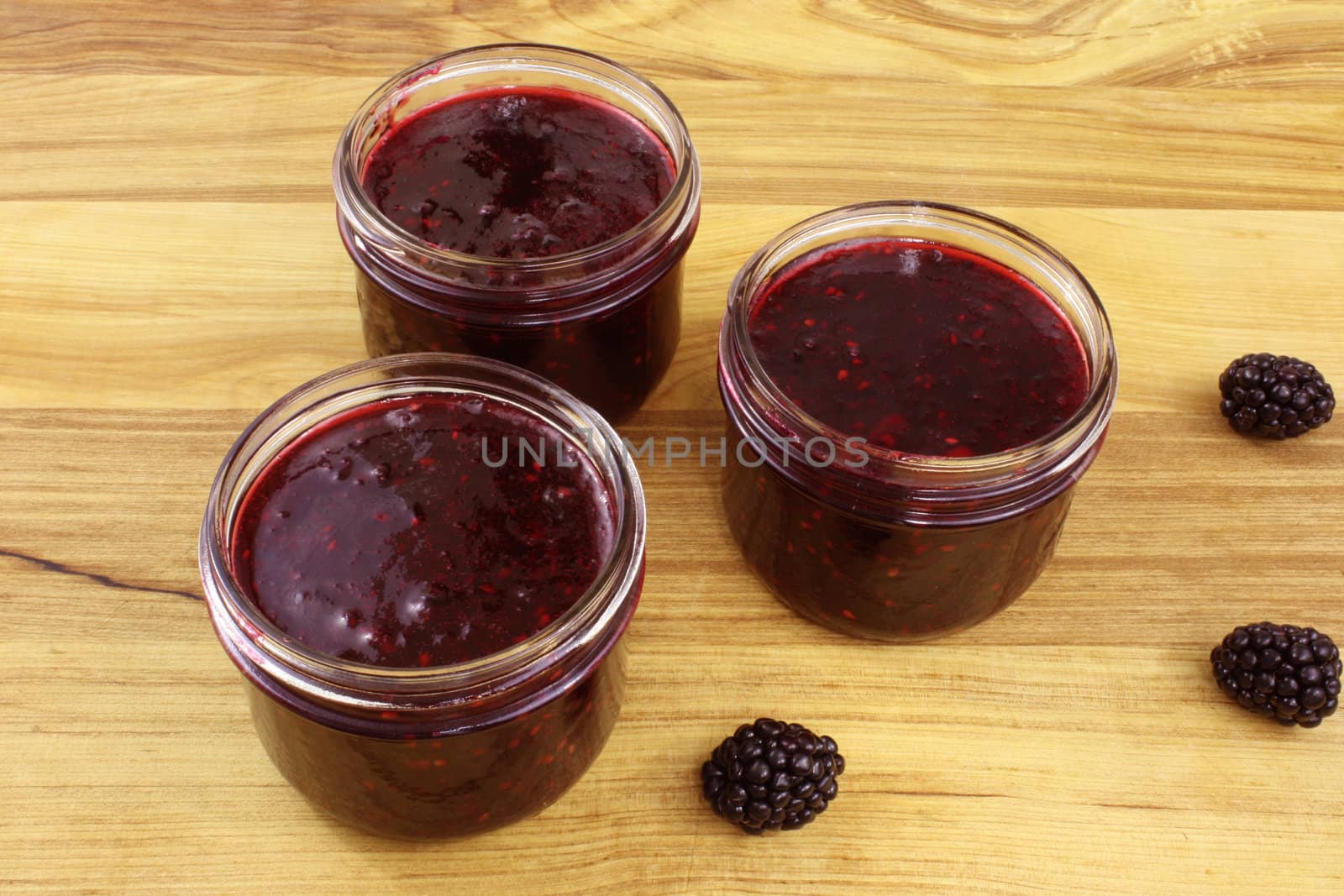 Three jars of home-made blackberry jam and three blackberries on a wooden countertop.