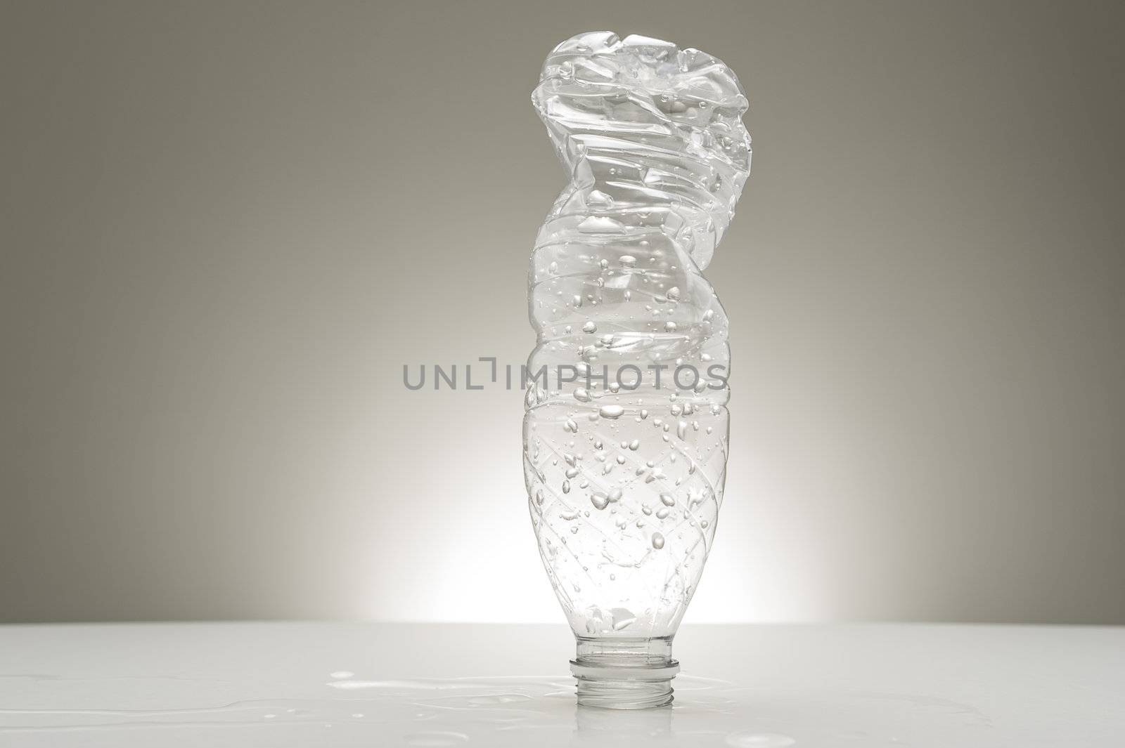 Empty recyclable plastic water bottle covered in water droplets and already partially twisted and compressed for recycling standing on its neck on a studio countertop
