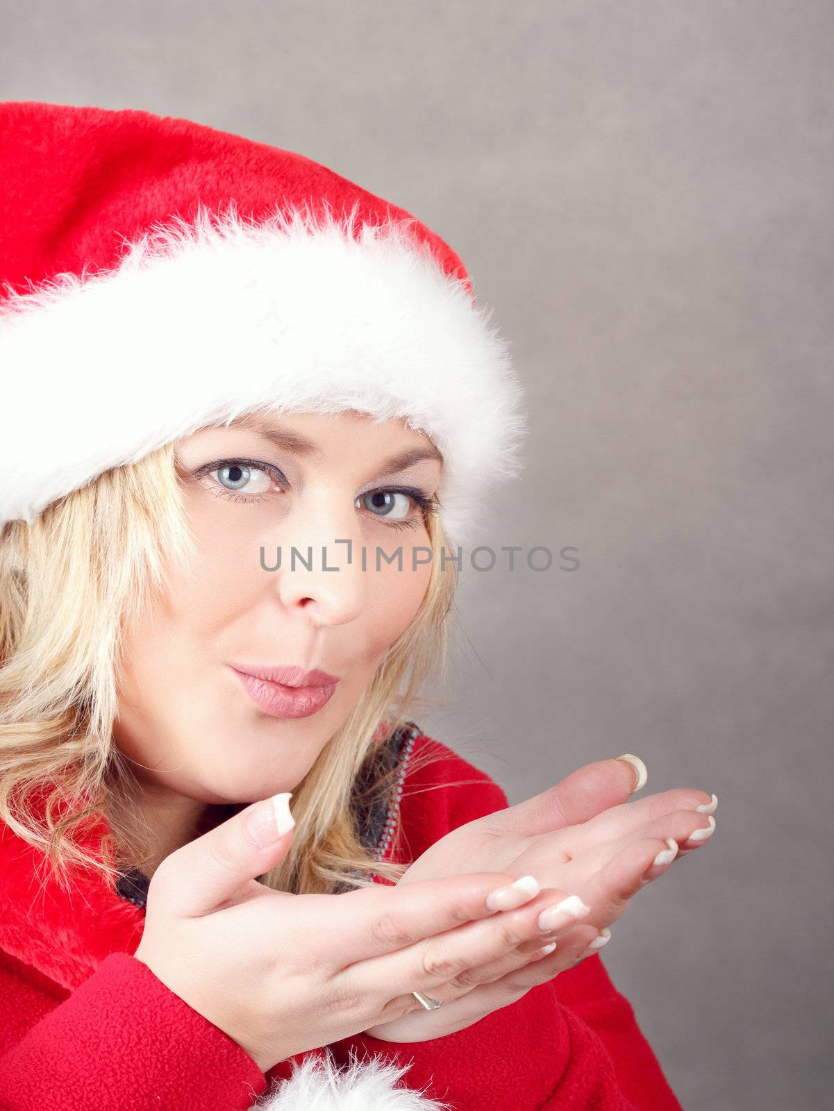Portrait of joyful pretty woman blowing in red santa claus hat on grey background