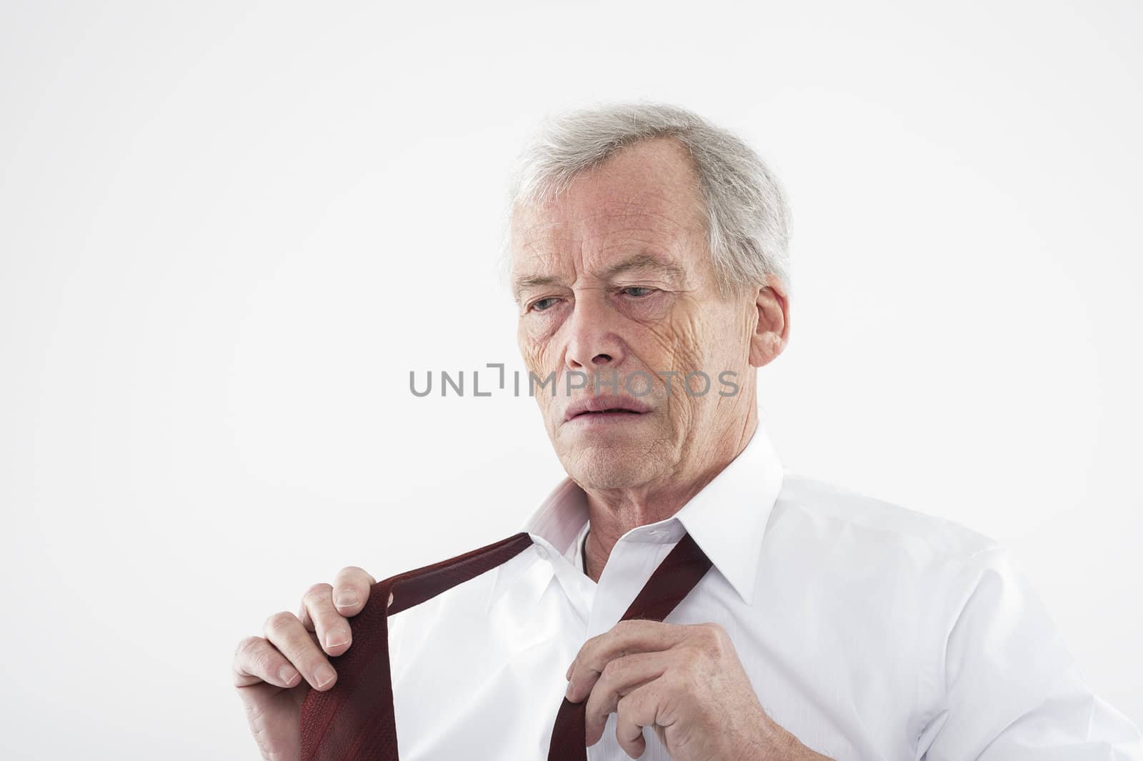 Serious elderly man with a pensive expression putting on his tie holding the two ends in his hands as he stands thinking, head and shoulders studio portrait
