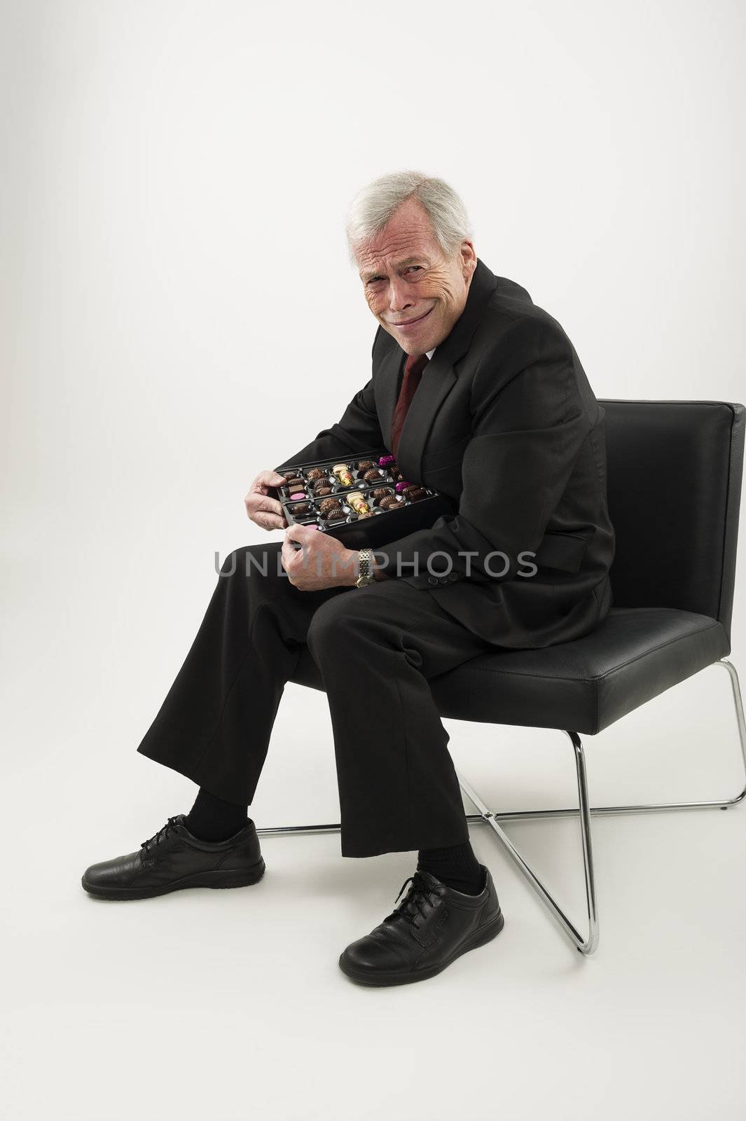 Studio portrait over white of an old man in a suit, sitting in a chair and protecting a large box of chocolates from anyone who may wish to share