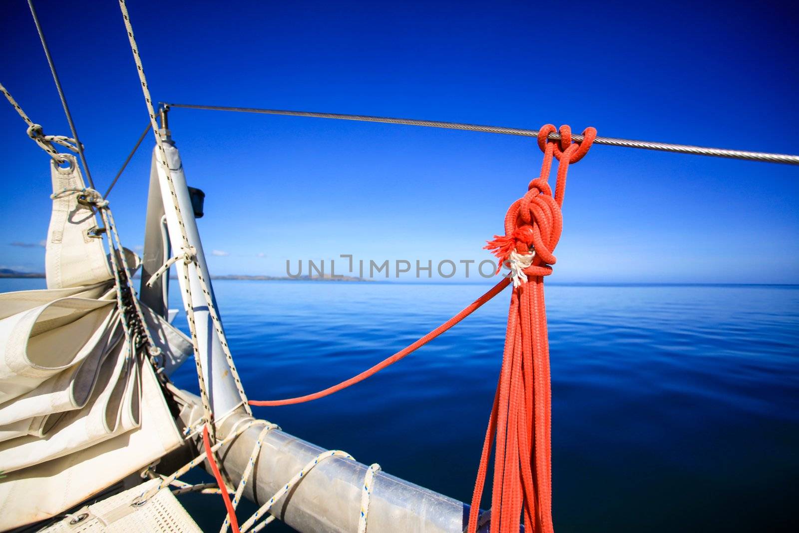 View of the bow of a sailing boat in calm blue sea with red rope riggings and white mast.