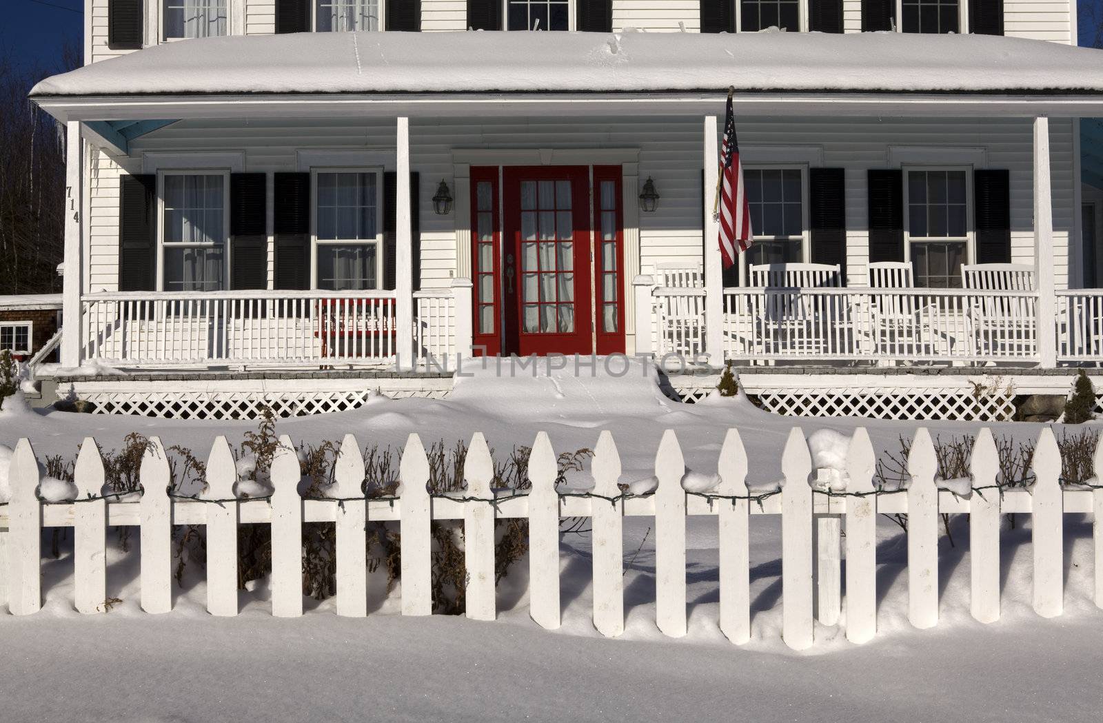 Front porch Of a Colonial style house covered in snow, red door, black shutters, white paint, white picket fence and American Flag.