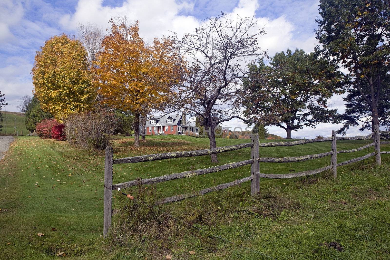 A vintage red brick cape style house in Autumn season surrounded by green lawn grass, colorul yellow, red and orange trees and a mossy, old split rail fence.