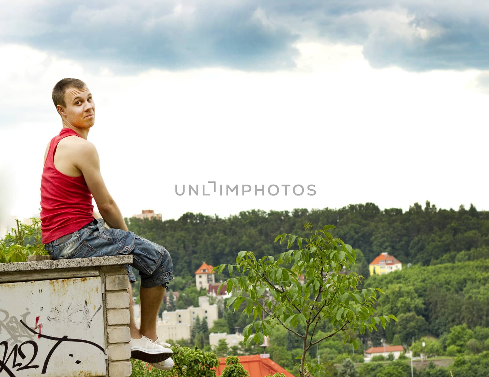 A guy sitting on a fence against panorama of foliage and roofs