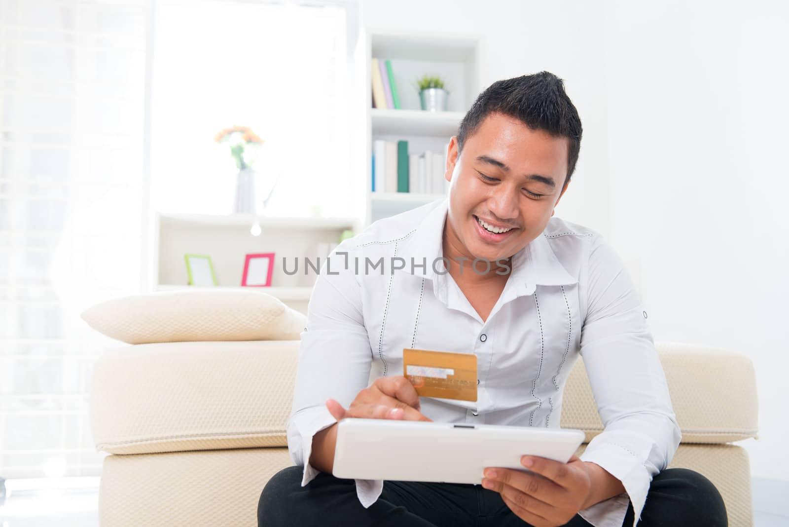 Happy Southeast Asian man online shopping, hands holding credit card and computer tab sitting at home.