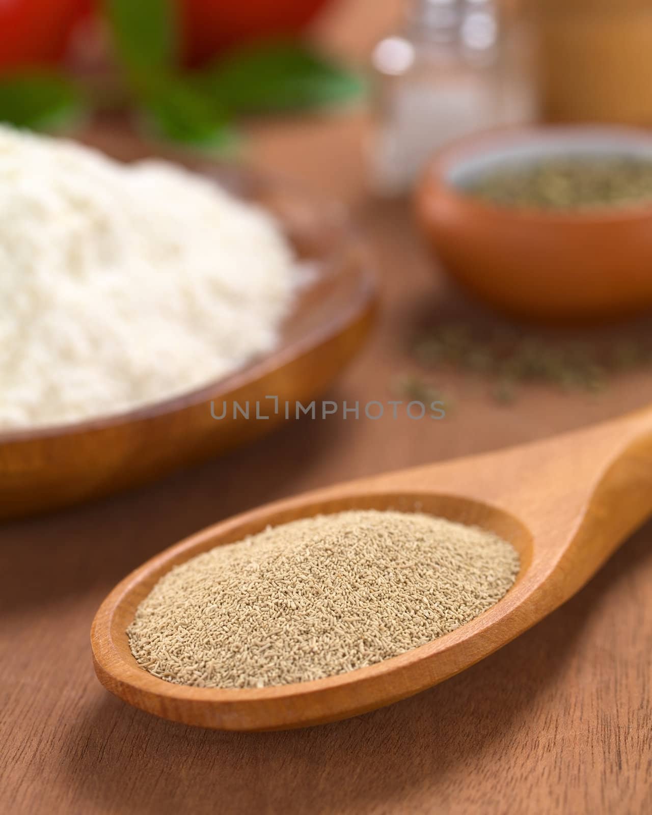 Basic ingredients of a pizza: Active dry yeast on wooden spoon with oregano, flour, salt, pepper, tomato and basil in the back (Selective Focus, Focus in the middle of the dry yeast)  