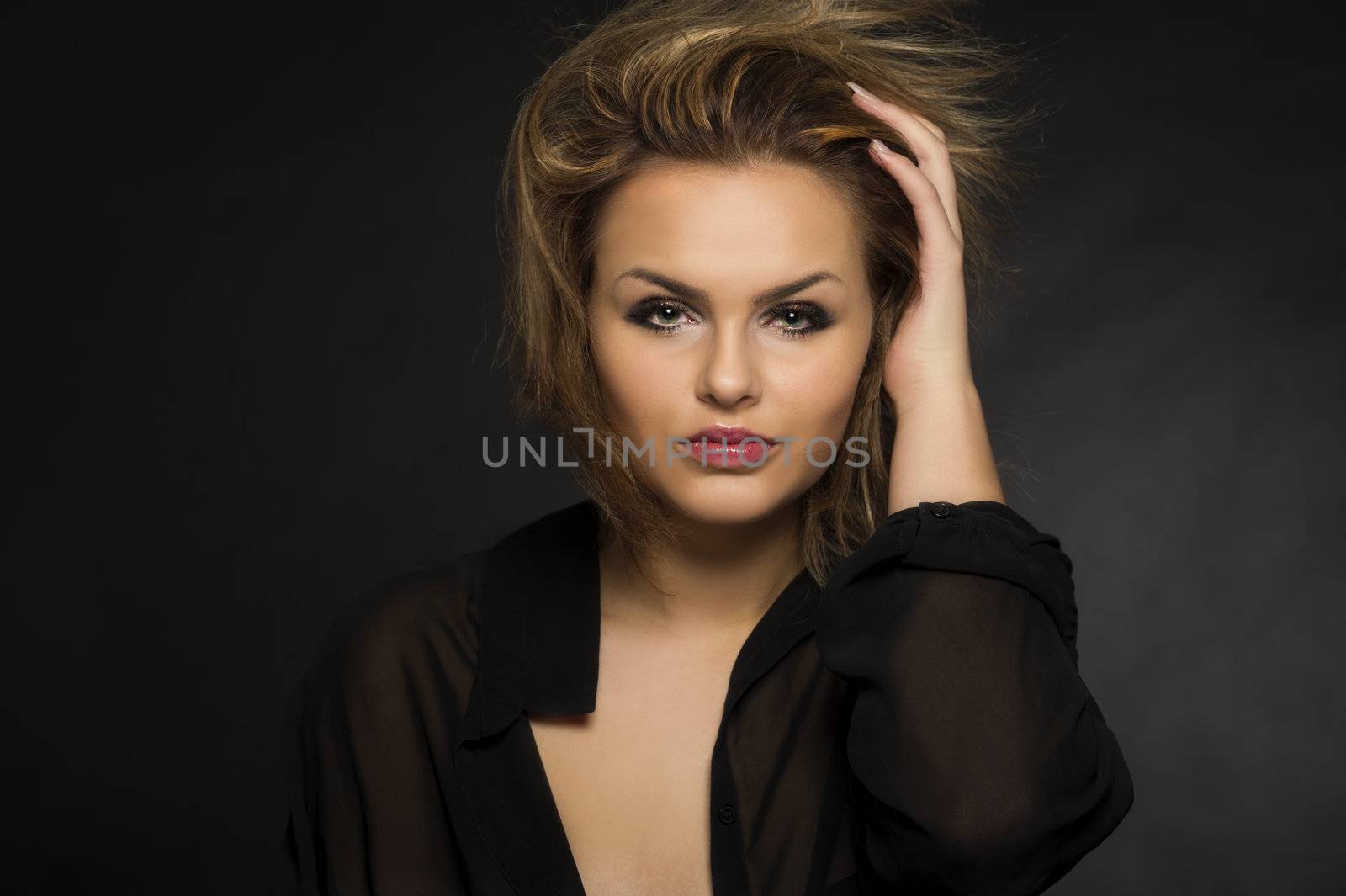 Beautiful woman with a windblown hairstyle holding her hand to her head as she looks at the camera, head and shoulders studio portrait on a dark background