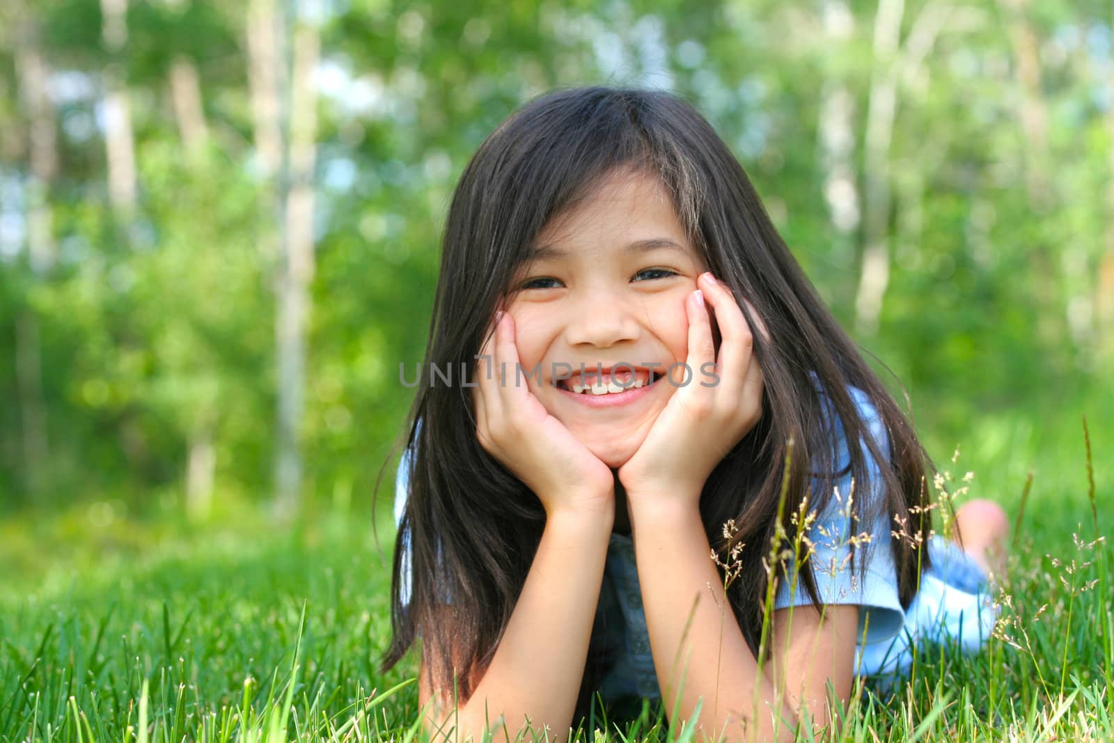 Child lying on grass with head on hands, smiling. Part asian, scandinavian background.