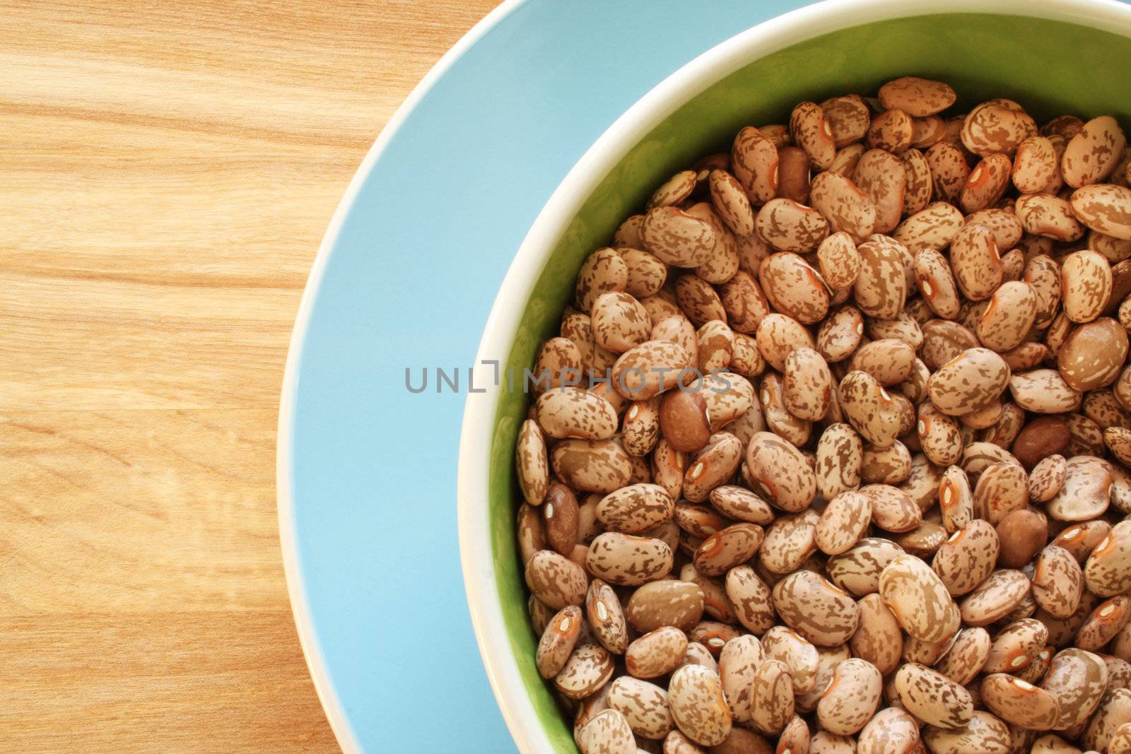 A close-up background or wallpaper image of heirloom dry Pinto beans in a green bowl, set upon a blue plate, all resting on a wooden counter top.