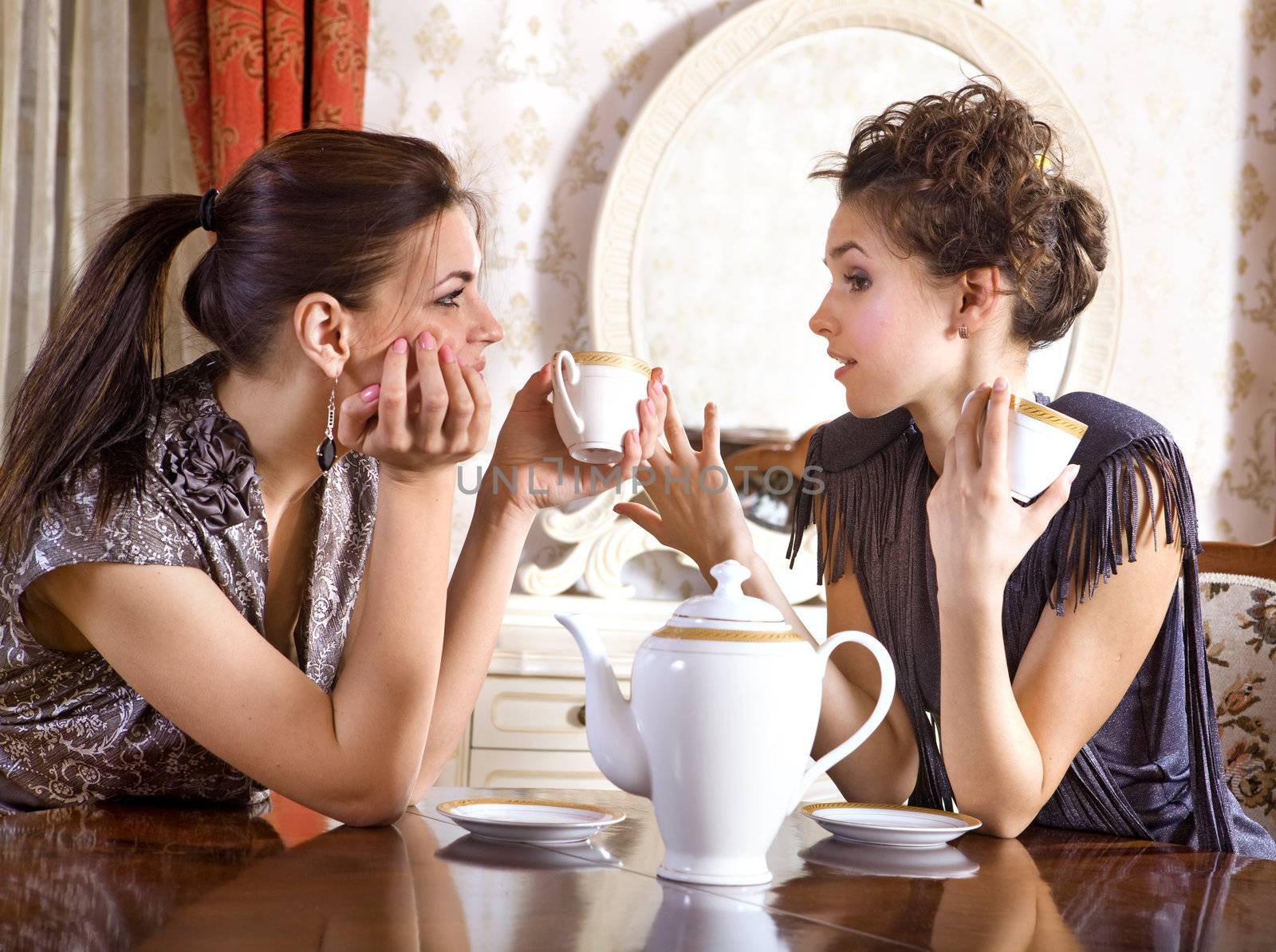 Two pretty girl-friends talk and drink tea in drawing room
