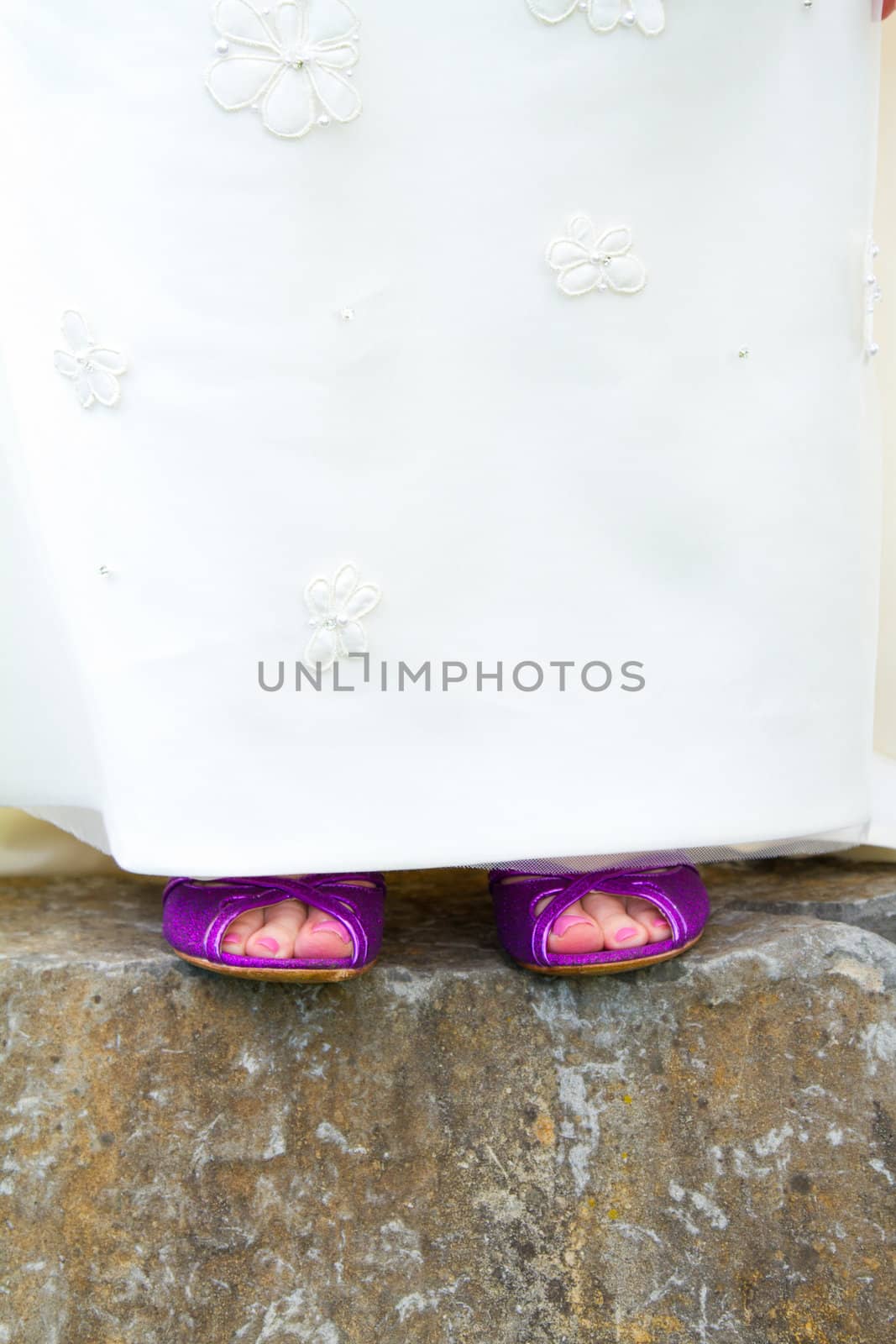 A beautiful bride in a white wedding dress stands outdoors with her pretty purple wedding shoes on her feet.